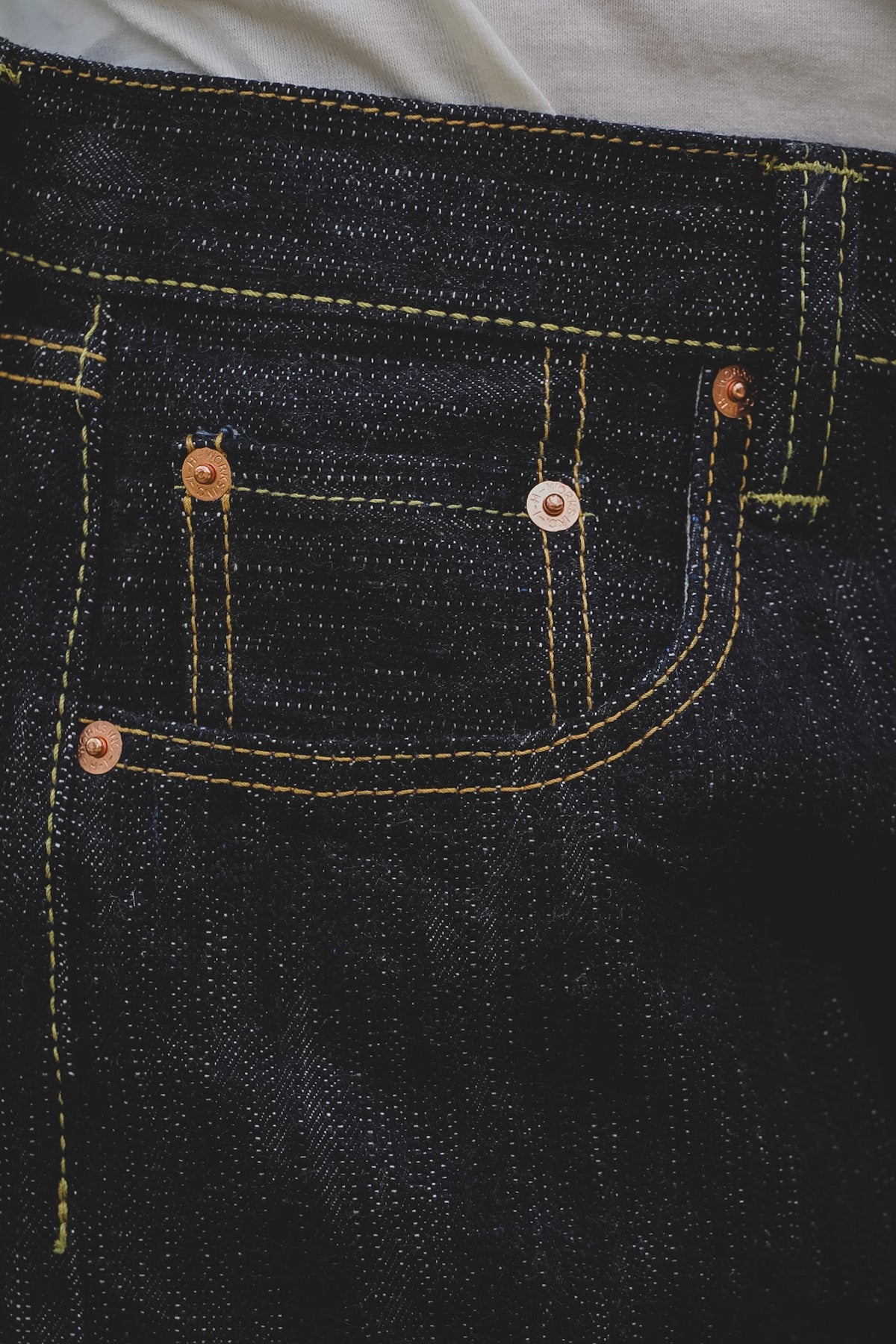 Close-up of dark indigo denim jeans showing the back pocket with yellow contrast stitching and copper-toned rivets.