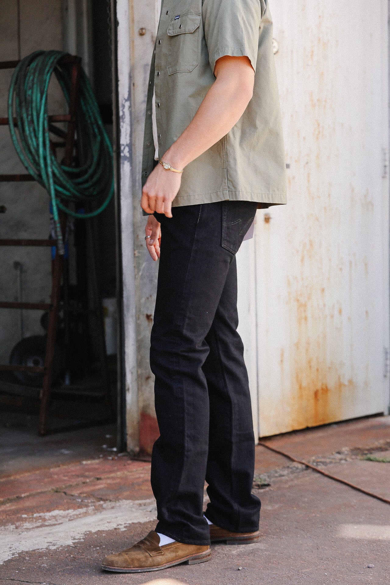 Person wearing black denim jeans, tan loafers, white socks, and an olive green short-sleeve shirt in a rustic outdoor setting