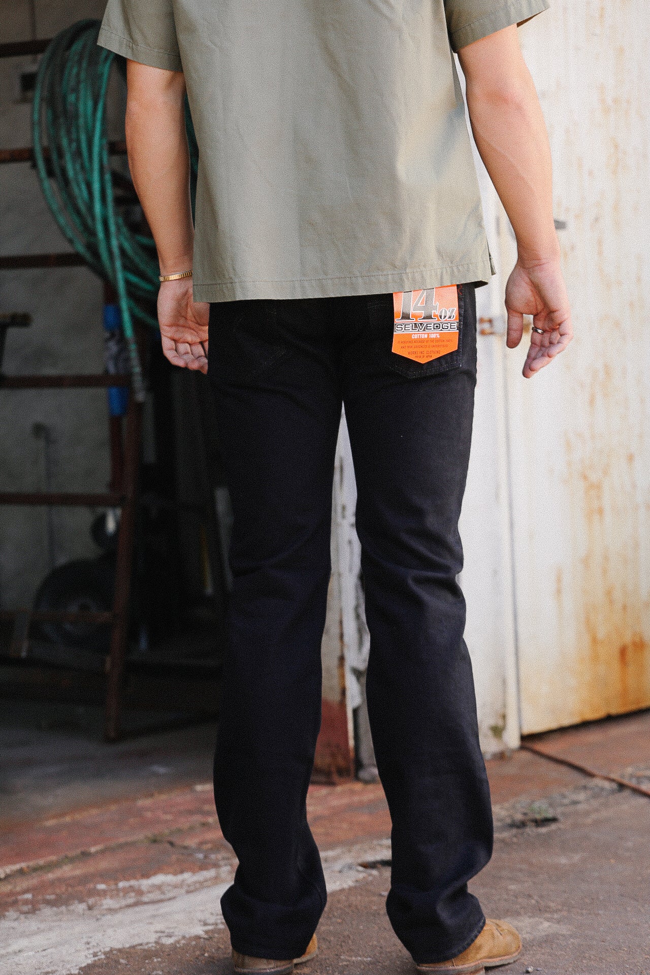 Man wearing black 14 oz selvedge denim jeans and olive green shirt in rustic industrial setting