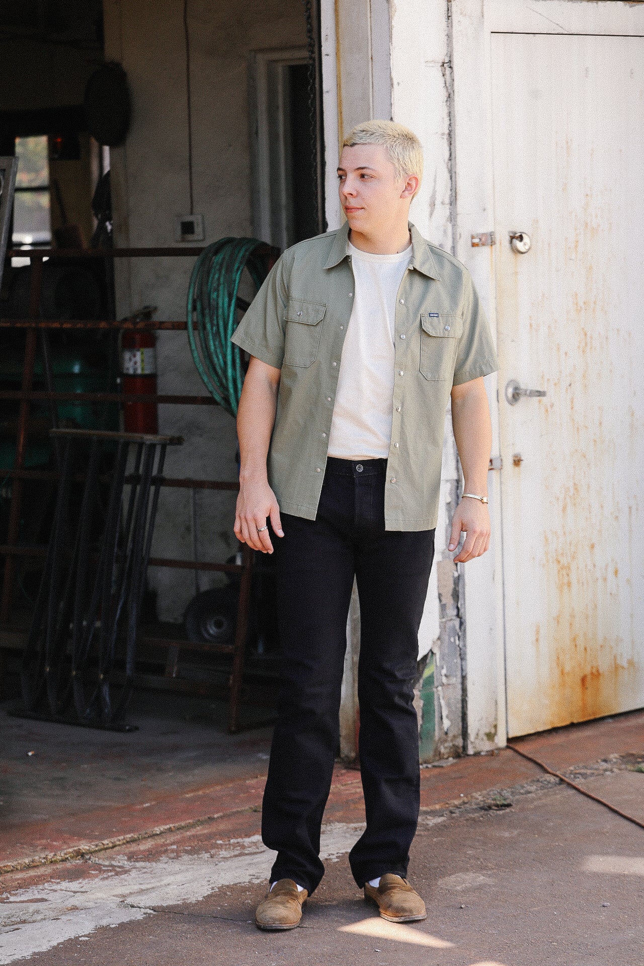 Young man wearing olive short-sleeve work shirt, white tee, black pants, and brown shoes in rustic workshop setting