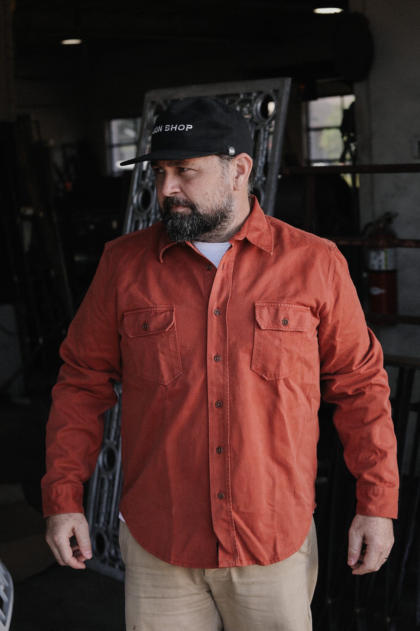 Man in rust-colored button-up shirt and black Iron Shop cap in workshop setting