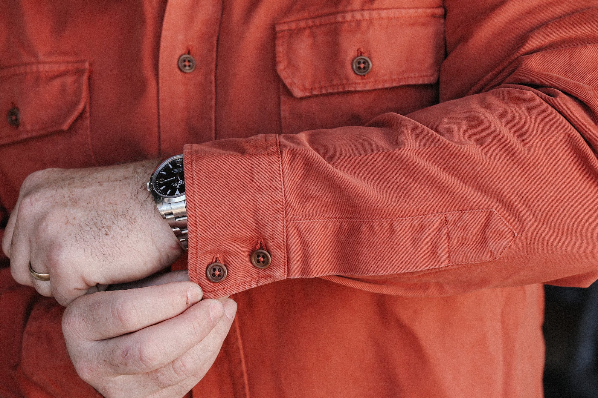 Close-up of a man adjusting the cuff of an orange work shirt with button details and wearing a stainless steel wristwatch