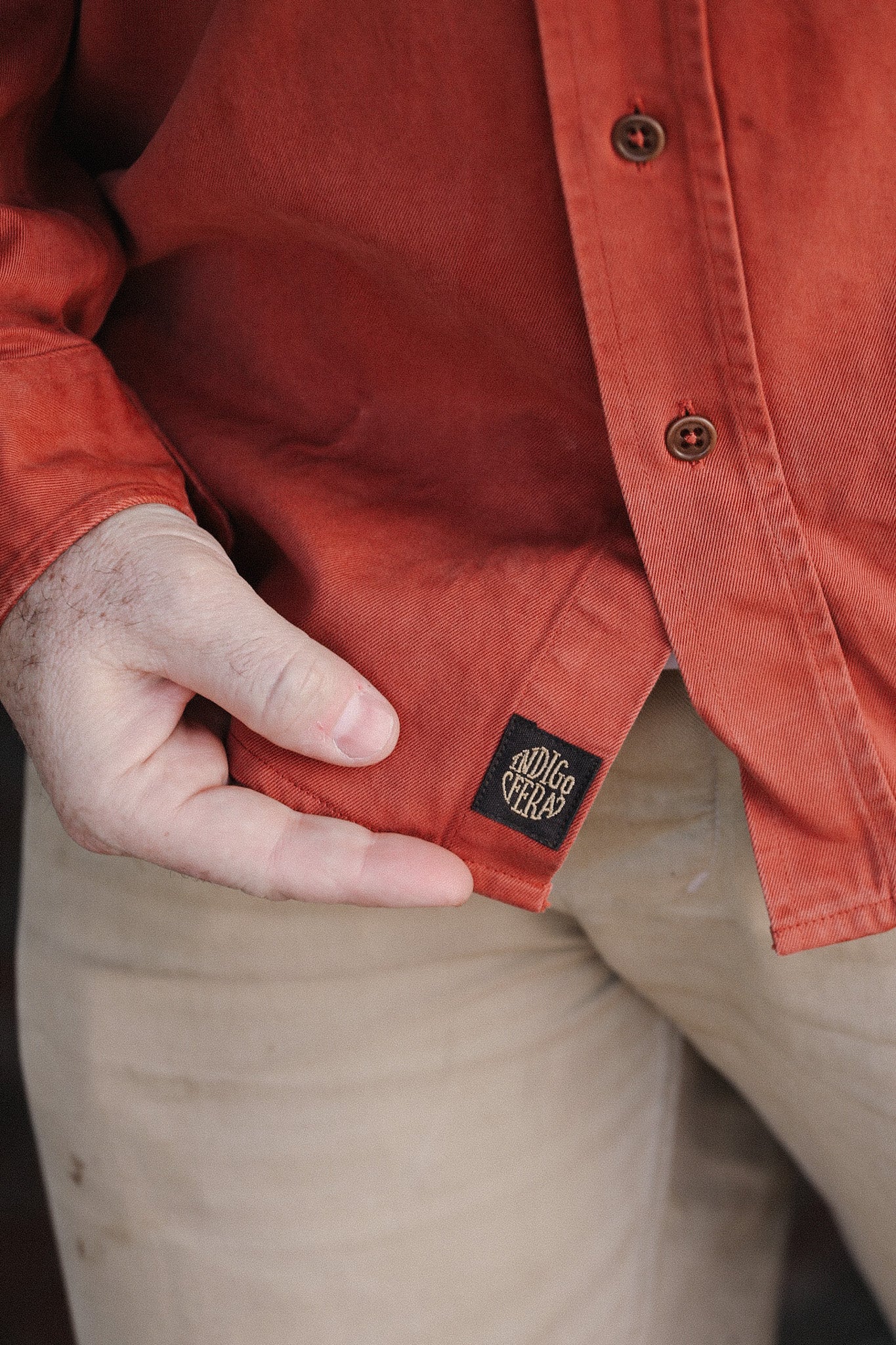 Close-up of person wearing rust-colored button-up shirt with visible Indigovera tag and beige pants