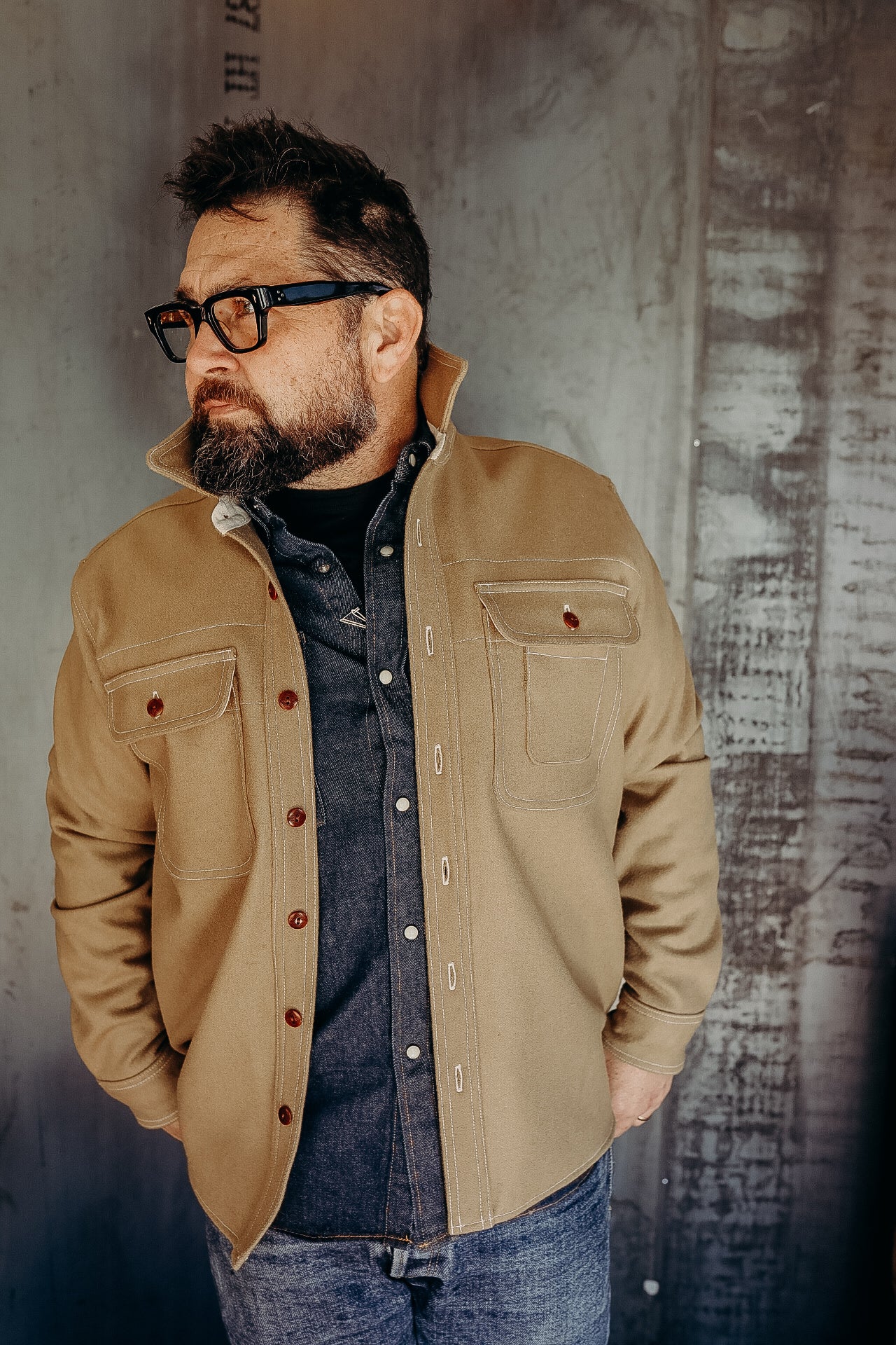 Man wearing tan work jacket over dark denim shirt standing against textured industrial wall