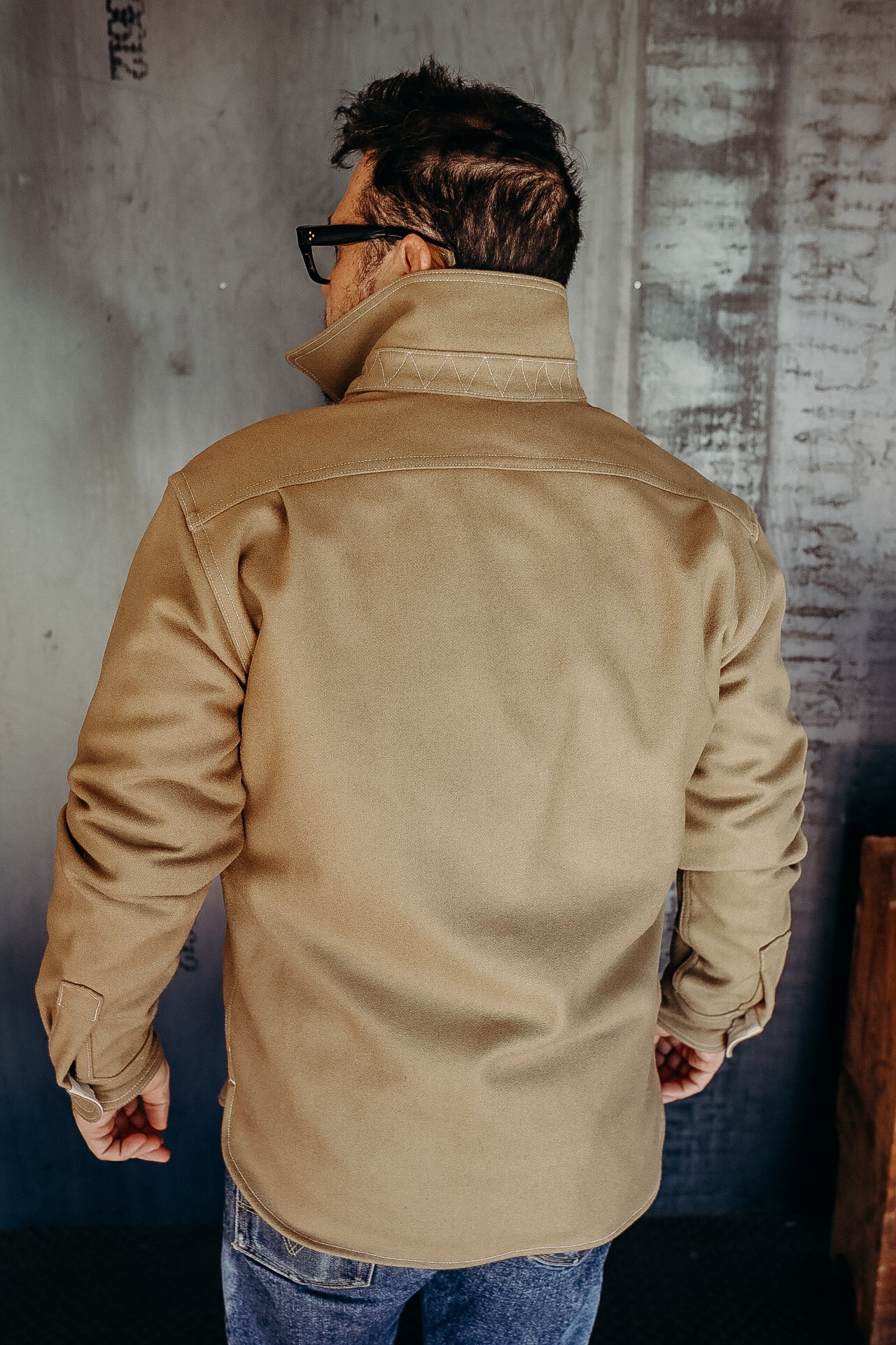 Man wearing tan rugged work jacket with raised collar and blue jeans in industrial background