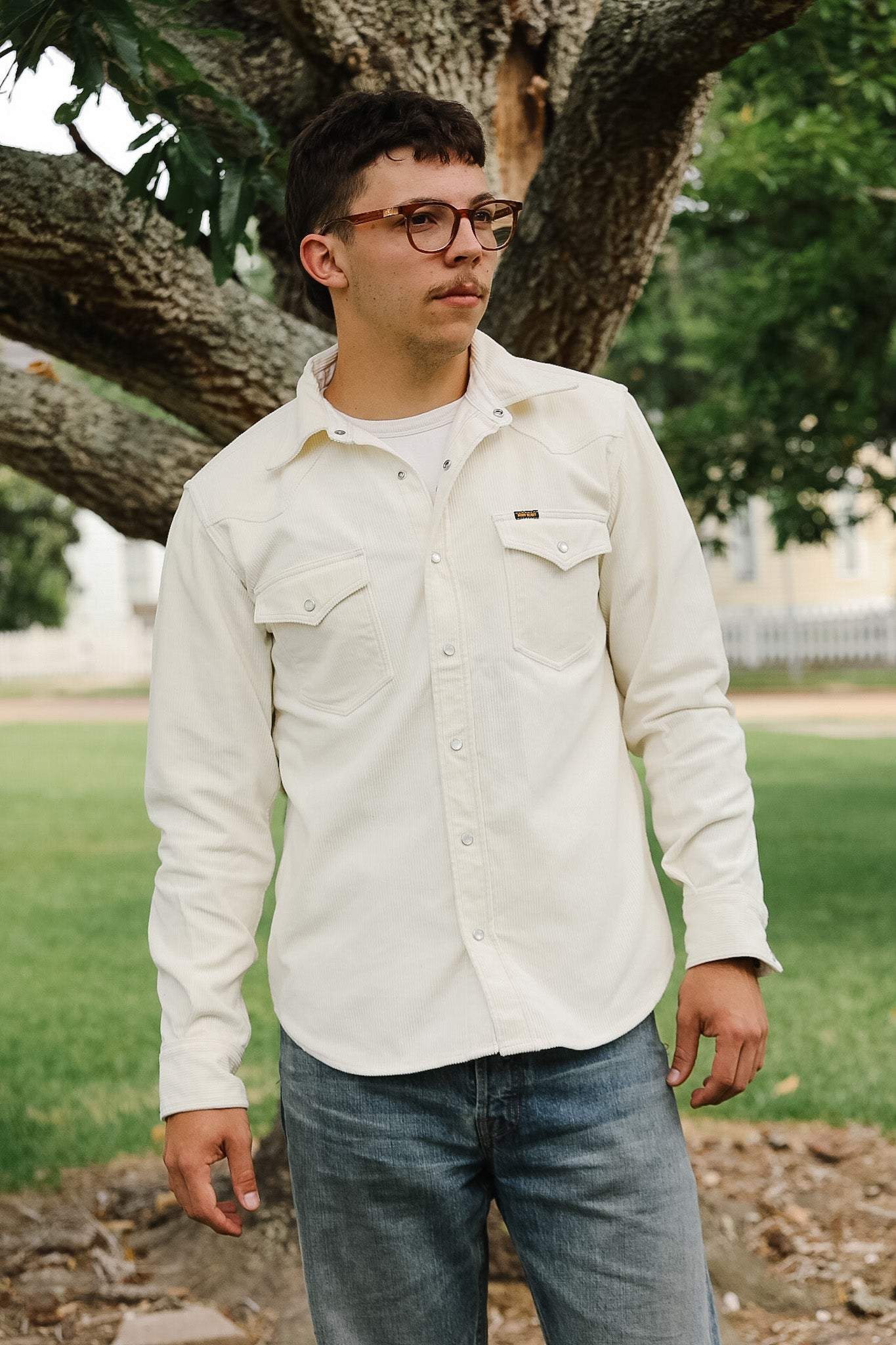 Man wearing a light-colored shirt and jeans standing outdoors with trees in the background