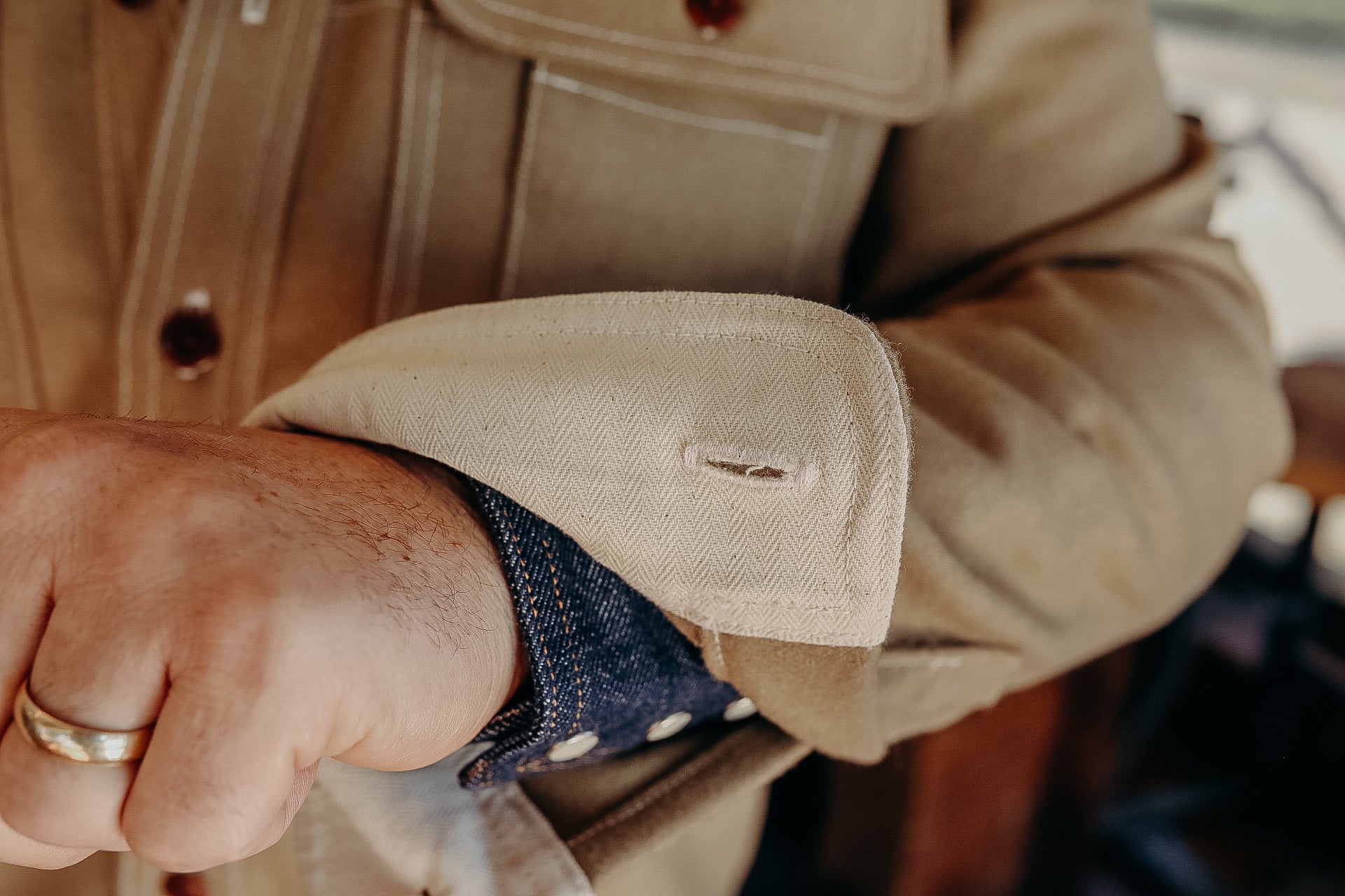 Close-up of a hand adjusting the cuff of a beige canvas jacket over a denim shirt sleeve, highlighting rugged workwear style