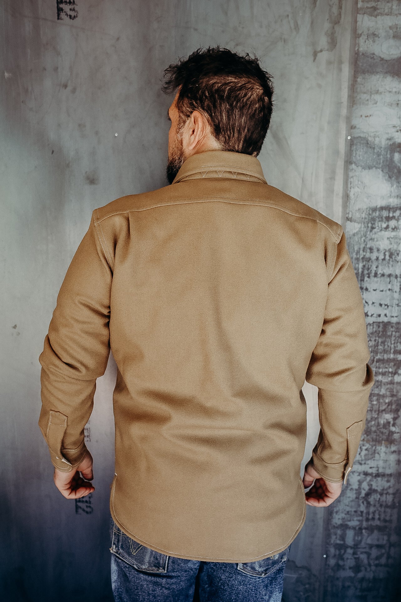 Man wearing a durable tan work shirt from Iron Shop Provisions, standing against textured wall