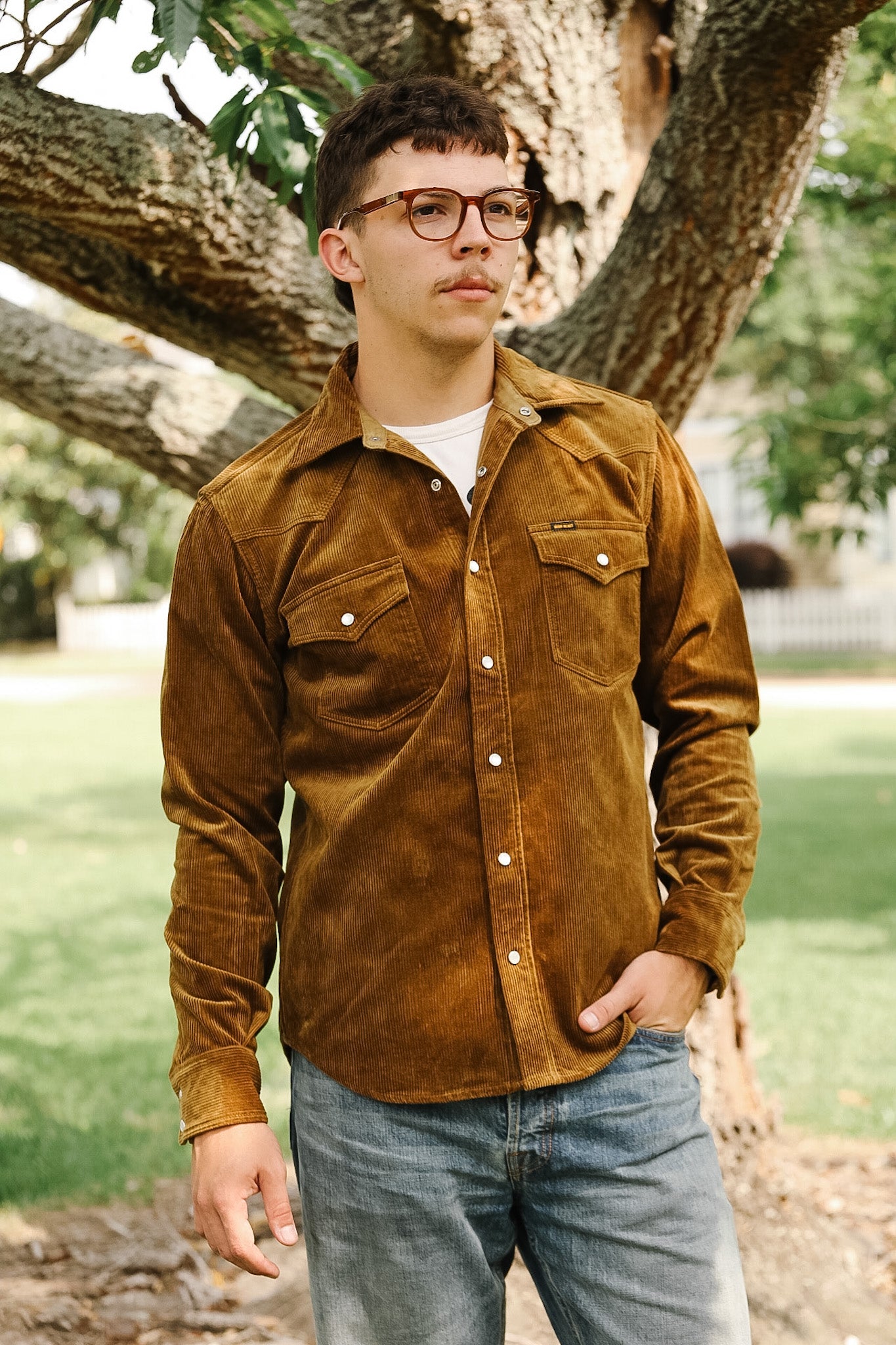 A man wearing a brown corduroy western shirt with a collar, button-up front, and cap, standing in front of a white background with a ladder in the background.