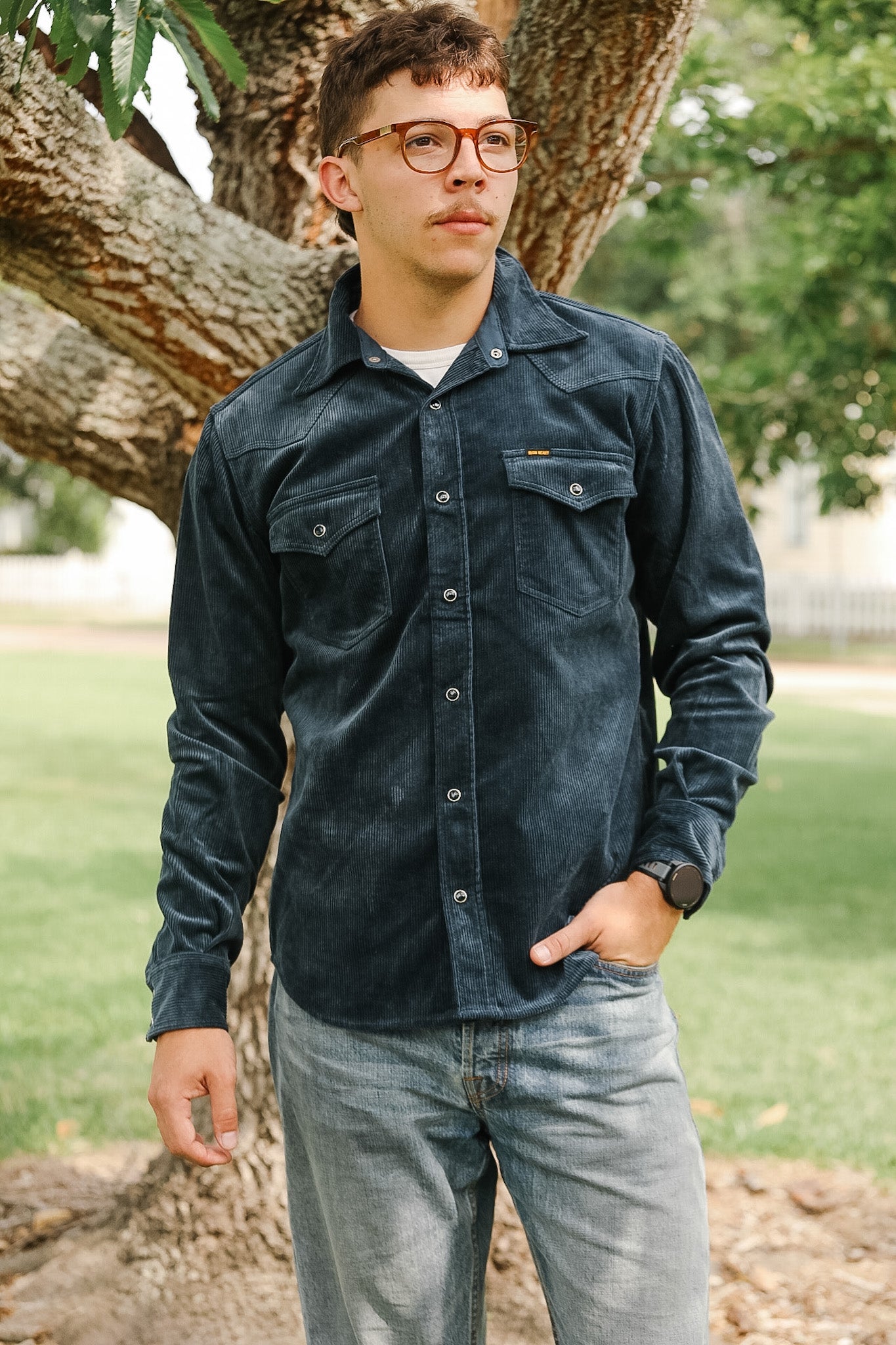 Man wearing a dark denim shirt and jeans standing outdoors near a tree.