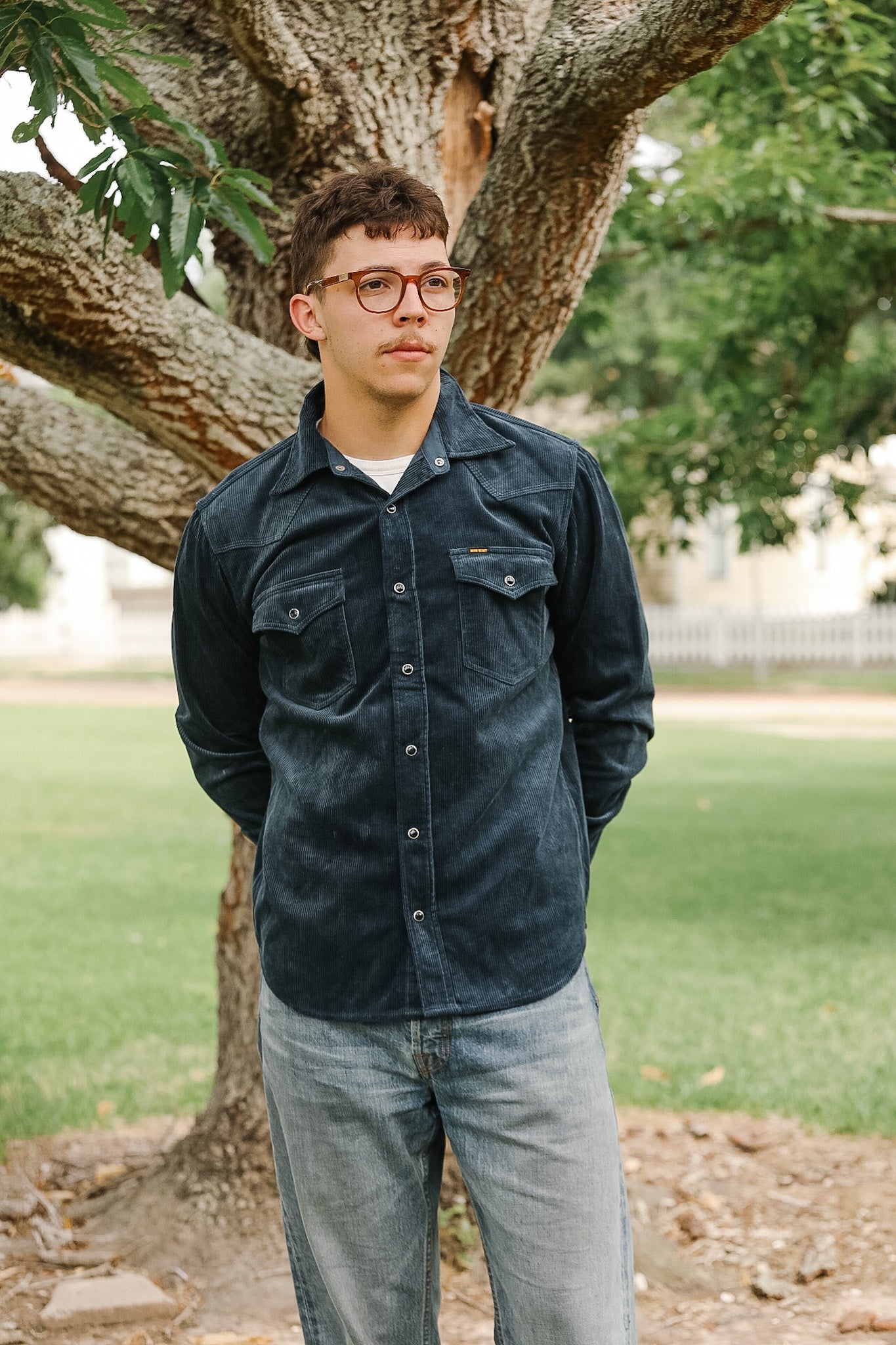 Person wearing a dark jacket and glasses standing under a tree in a park.