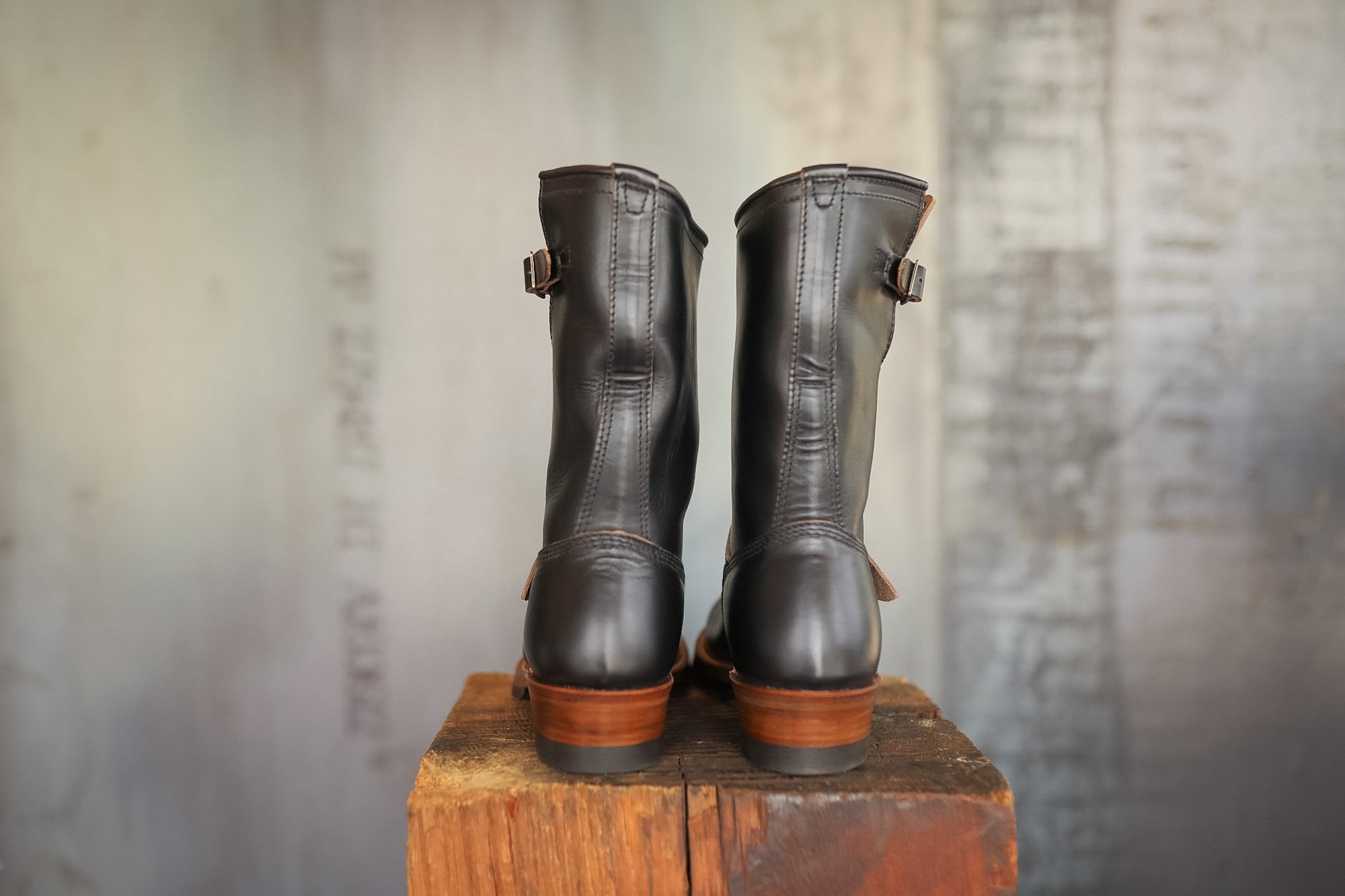 Pair of black leather boots with brown soles on a wooden block against a textured wall.