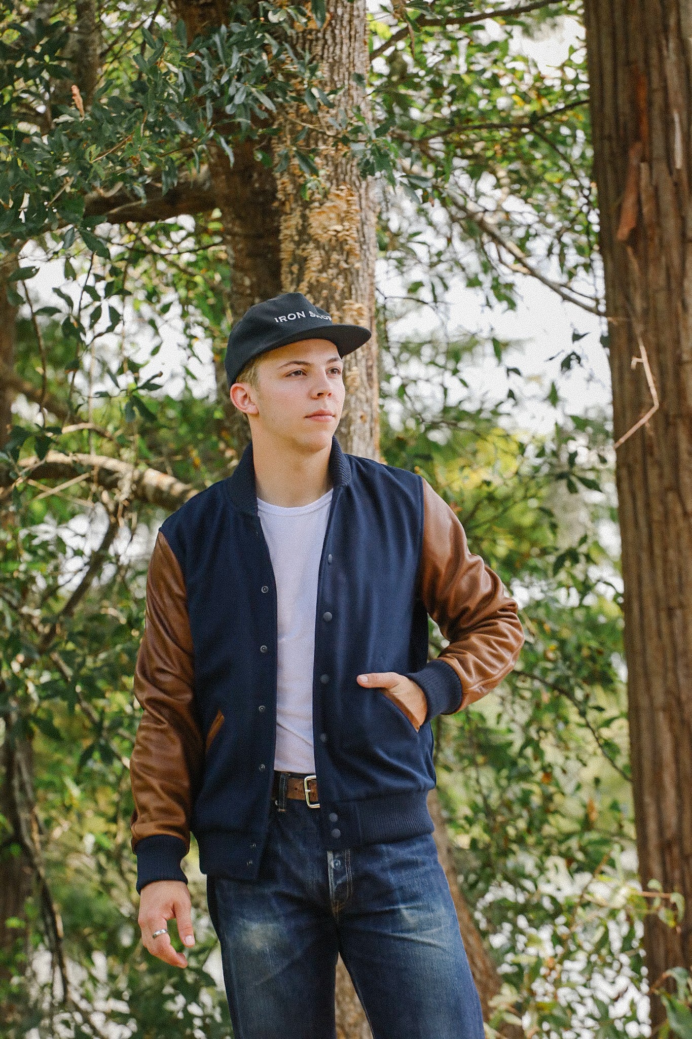 Young man in a navy blue and brown leather sleeve jacket and black Iron Shop Provisions cap standing outdoors near trees