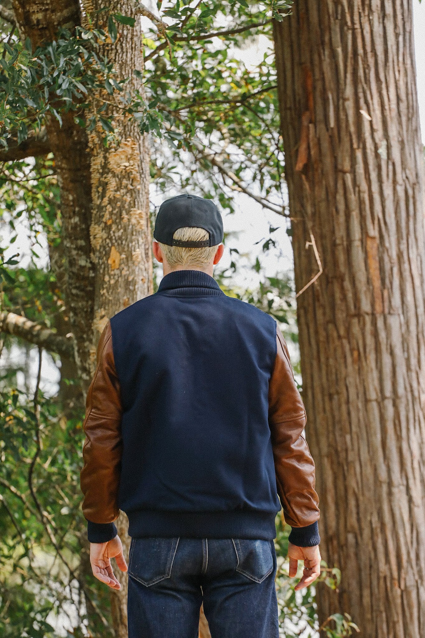 Person in navy varsity jacket with brown leather sleeves and black cap standing in forest