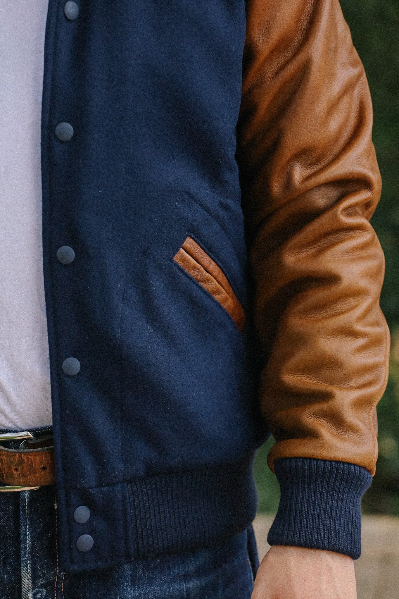 Close-up of a man wearing a navy and brown leather sleeve varsity jacket with ribbed cuffs and snap buttons