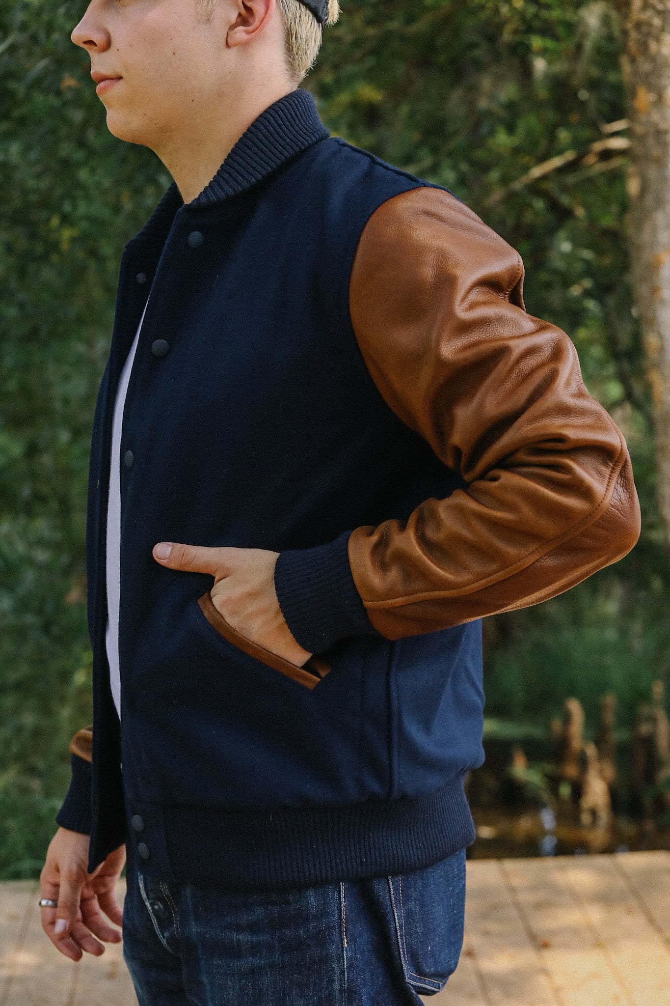 Man wearing navy blue and brown leather sleeve varsity jacket standing outdoors near greenery
