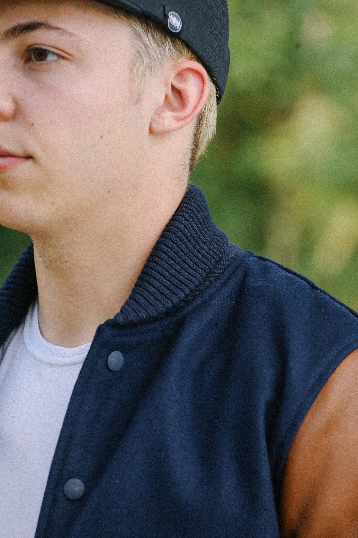 Close-up of man wearing navy blue varsity jacket with brown leather sleeves and black cap outdoors