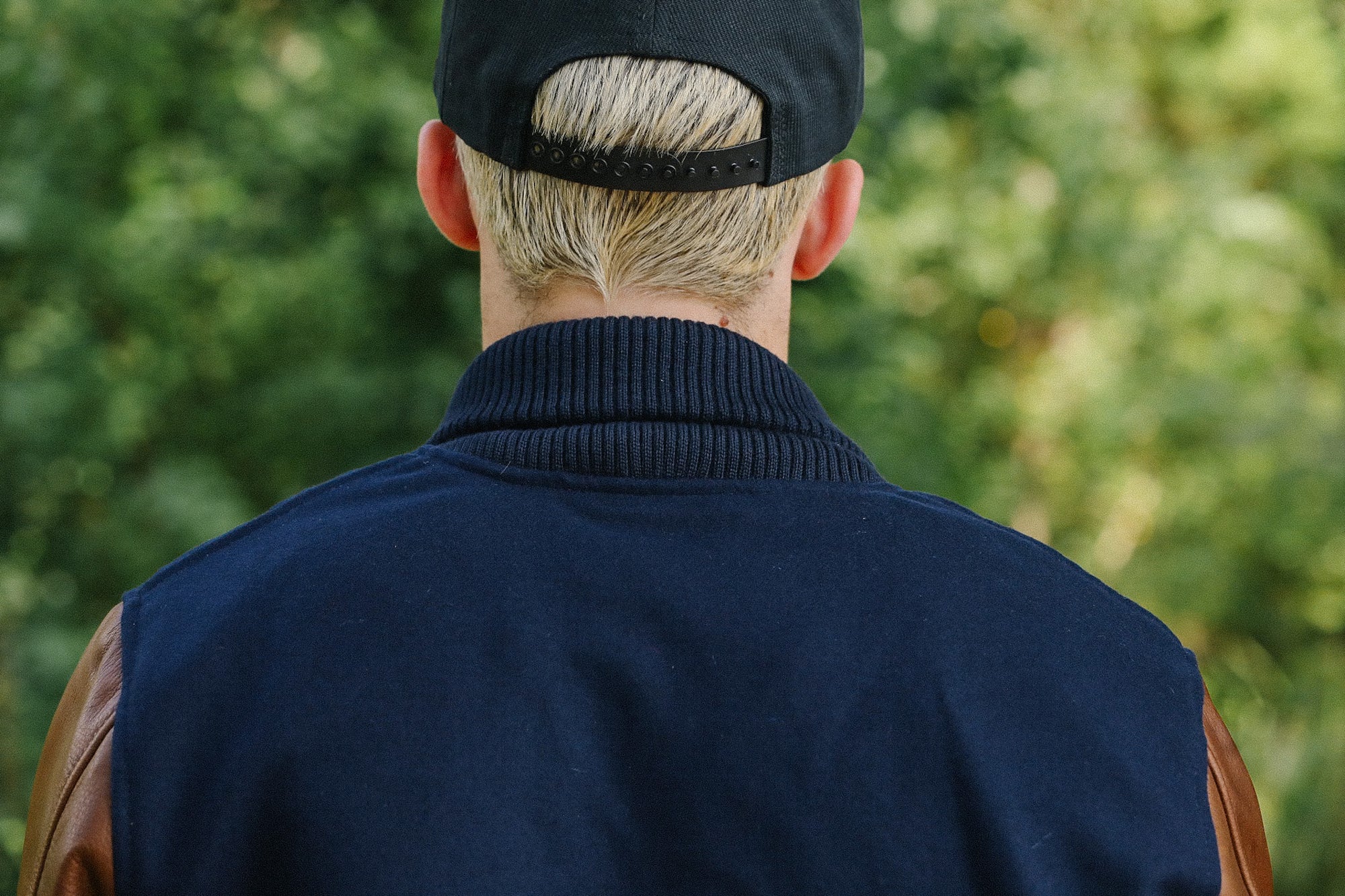 Man with blonde hair wearing a black snapback cap and navy jacket with brown leather sleeves outdoors