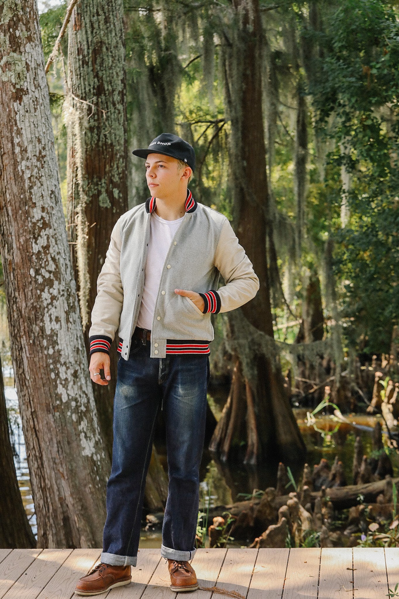Young man wearing Iron Shop Provisions jacket and cap standing on wooden deck in forested wetland