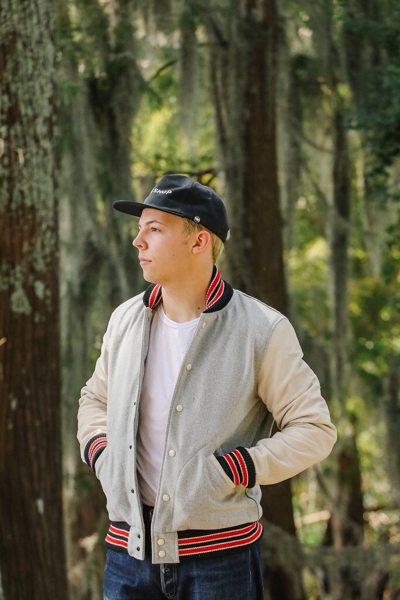 Young man wearing grey and beige varsity jacket with red striped trim and black Iron Shop cap in forest