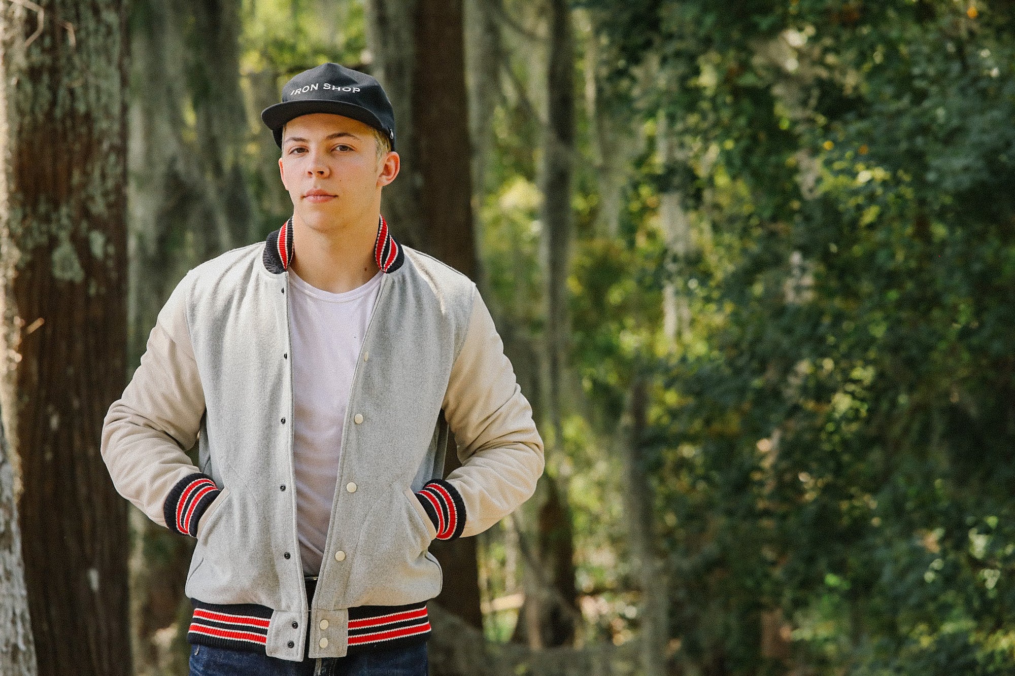Young man wearing Iron Shop black cap and vintage varsity jacket in wooded park setting