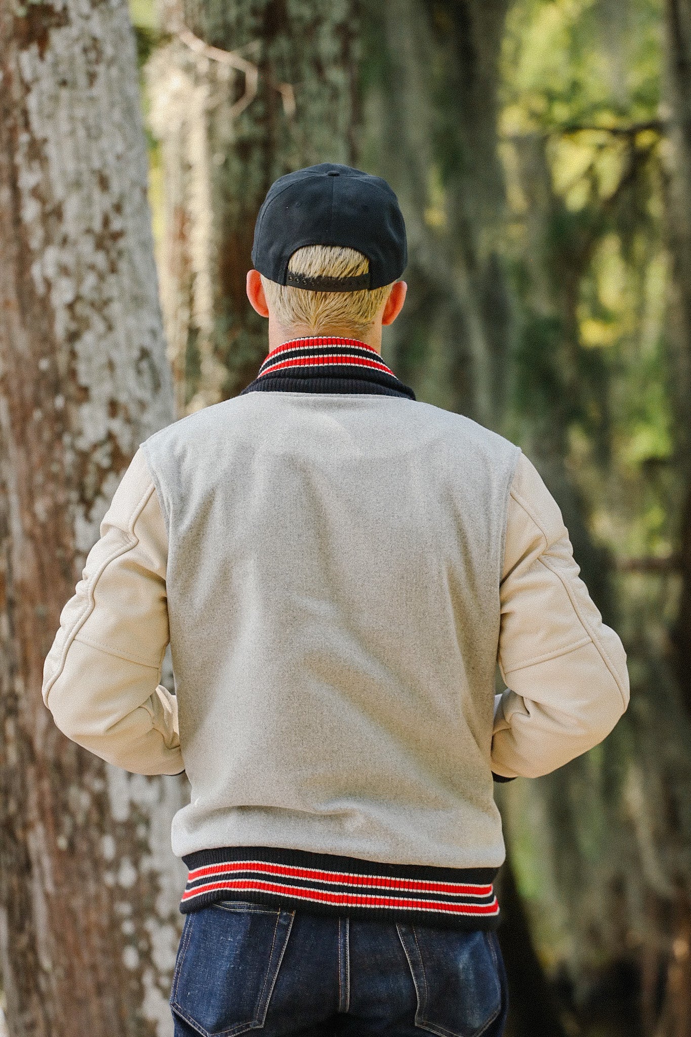 Man wearing beige and gray varsity jacket with red and black striped collar and a black cap outdoors