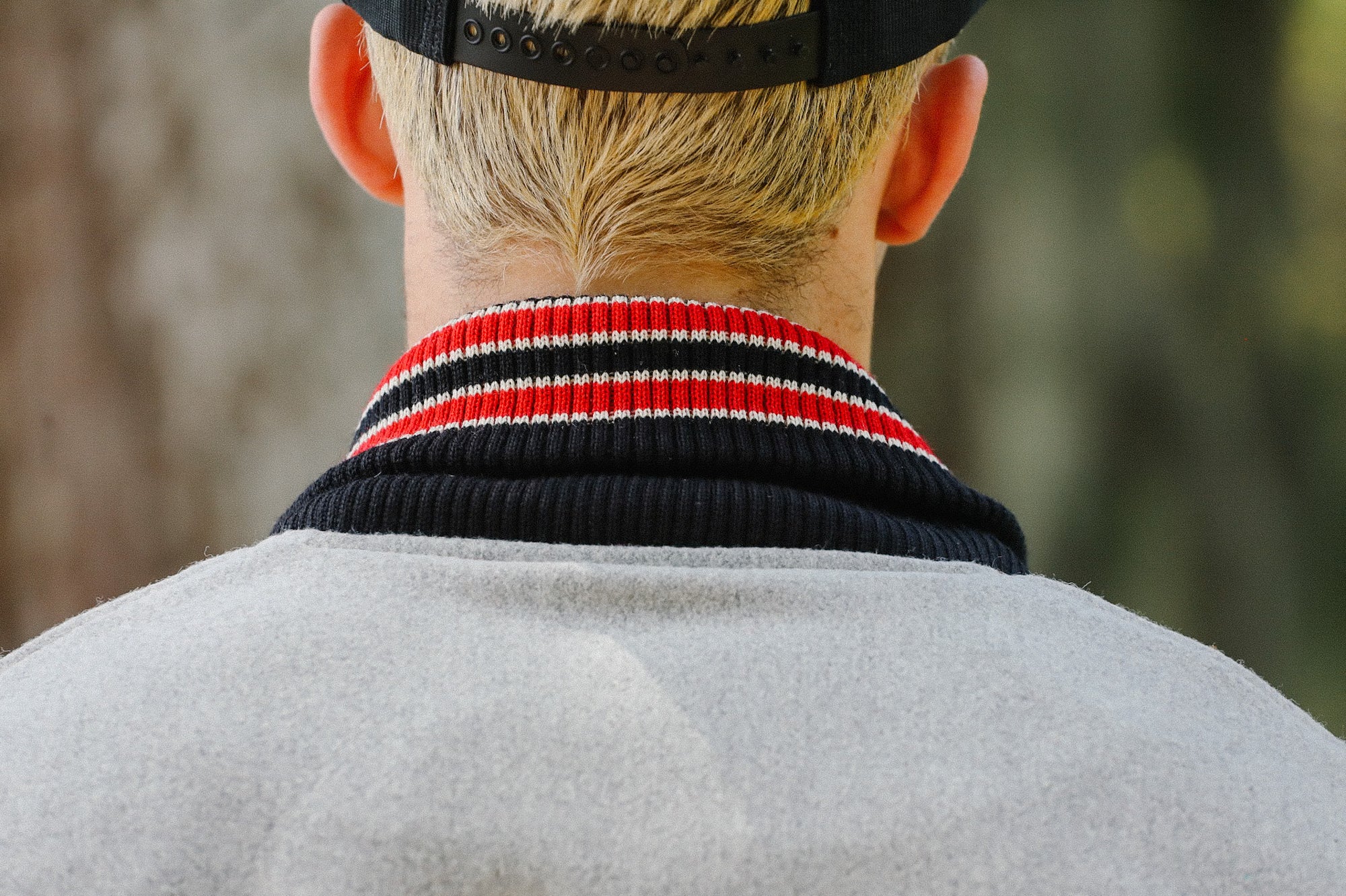 Back view of person with blond hair wearing black cap and gray jacket with red and black striped collar