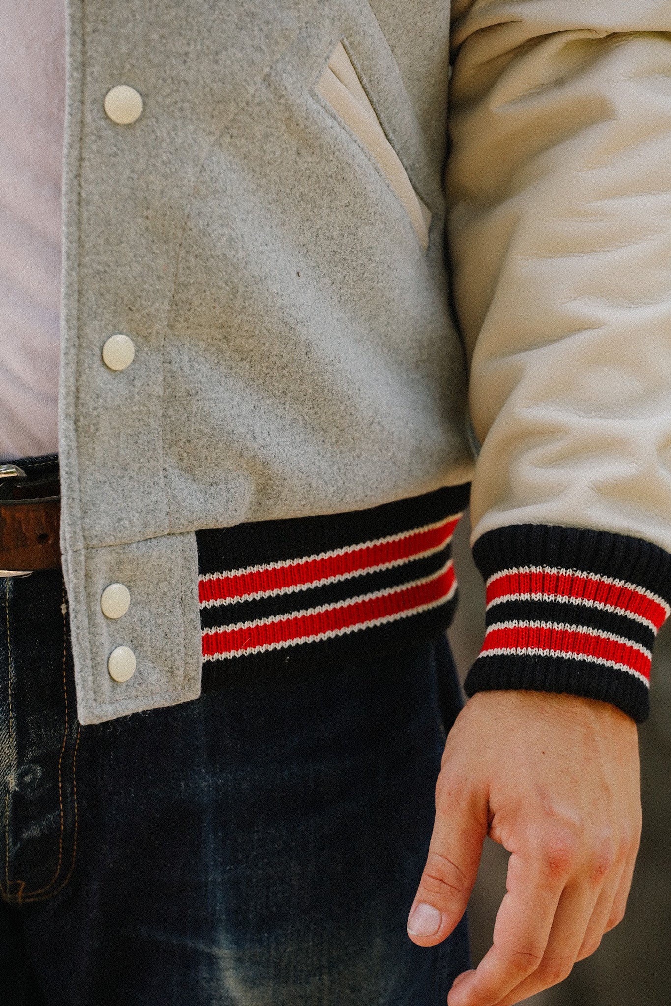 Close-up of a person wearing a grey and cream varsity jacket with red and black striped cuffs and blue denim jeans