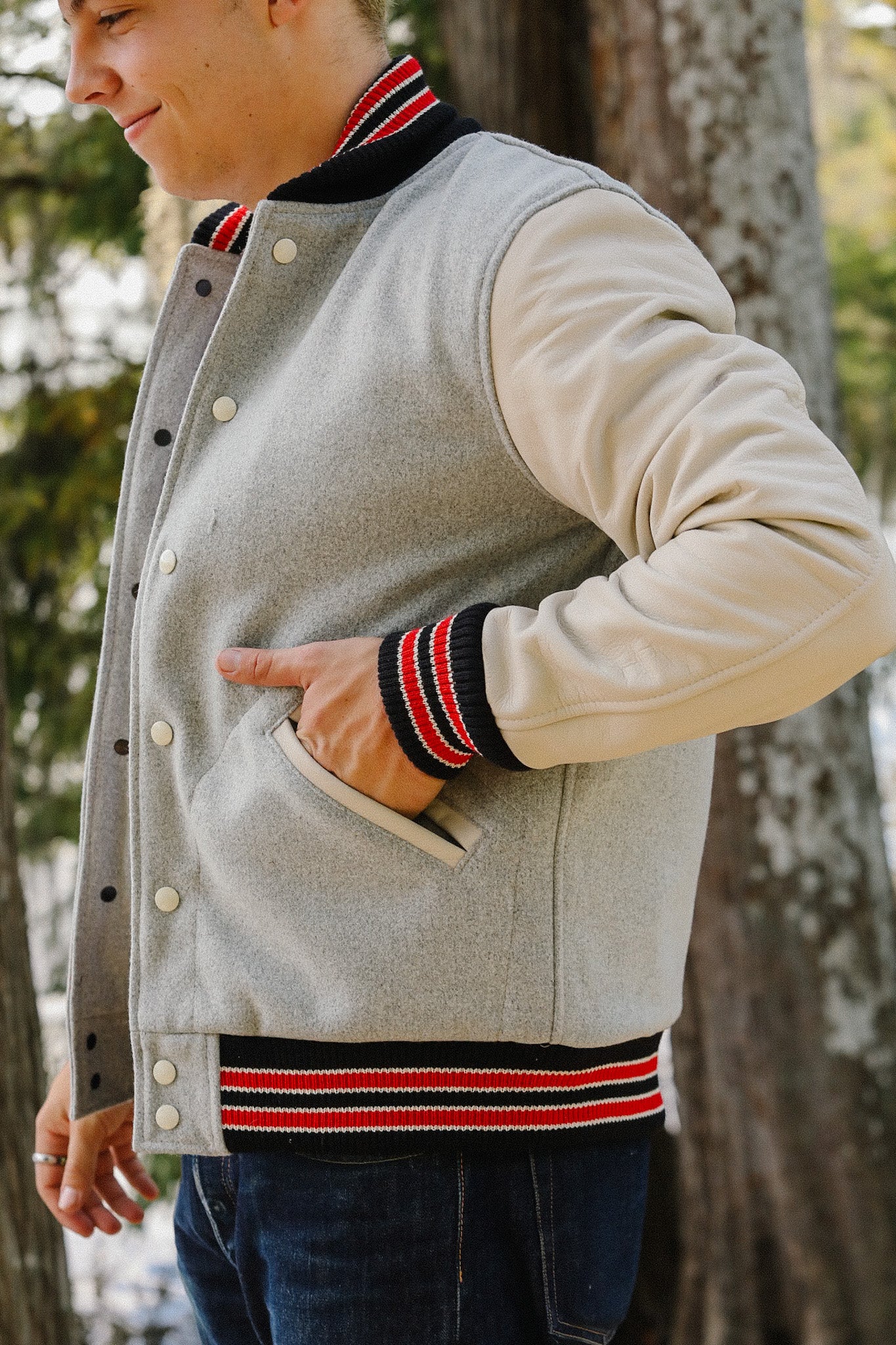 Man wearing gray varsity jacket with cream sleeves and red black striped cuffs, outdoors near trees