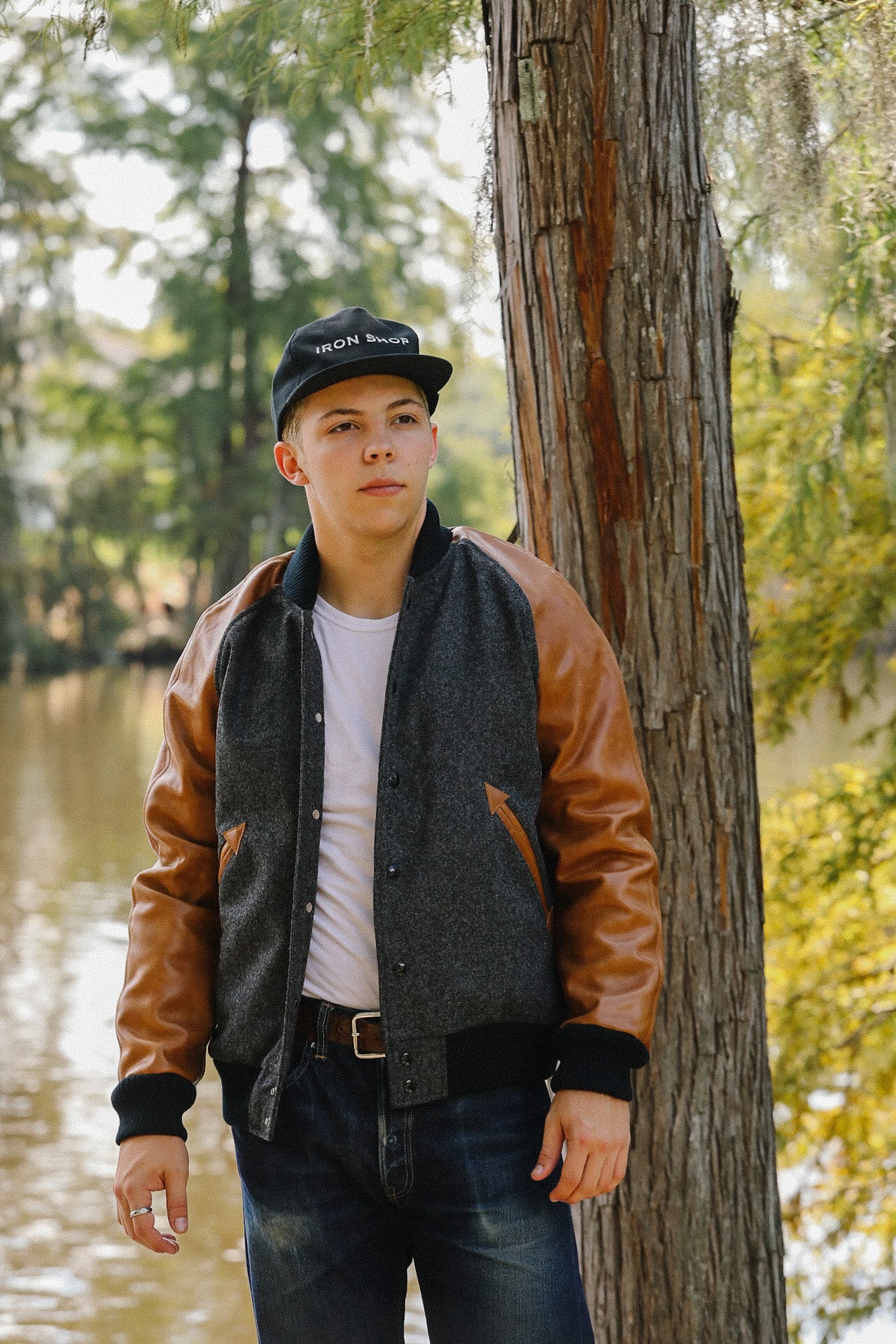 Young man wearing Iron Shop cap and leather-sleeve jacket standing by tree near lake in outdoor setting