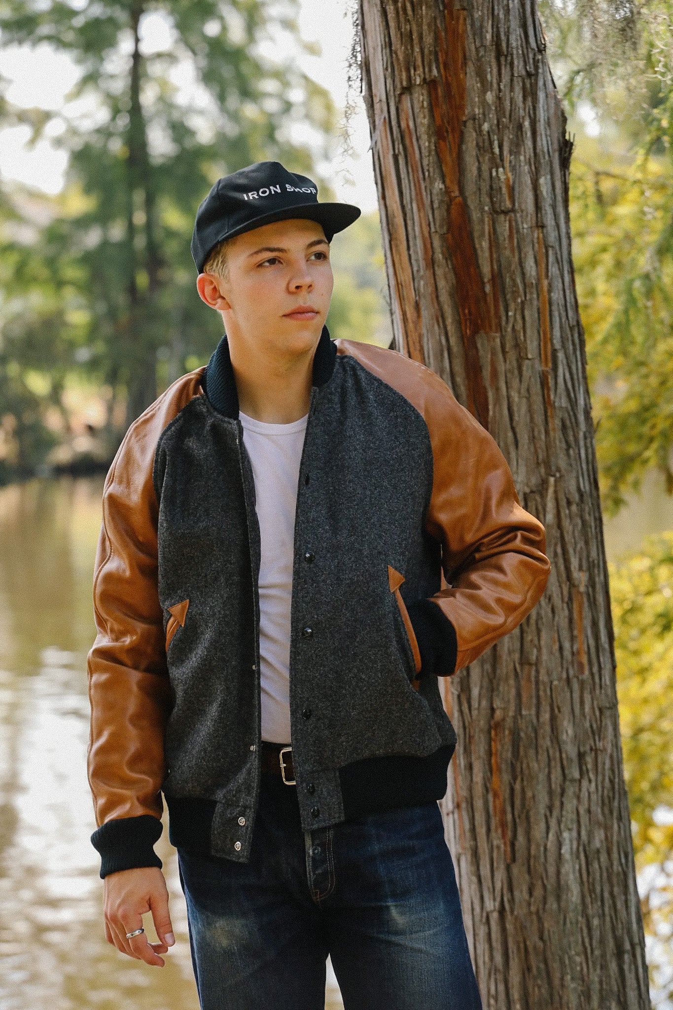 Young man wearing an Iron Shop cap and brown leather sleeve jacket by a tree near water