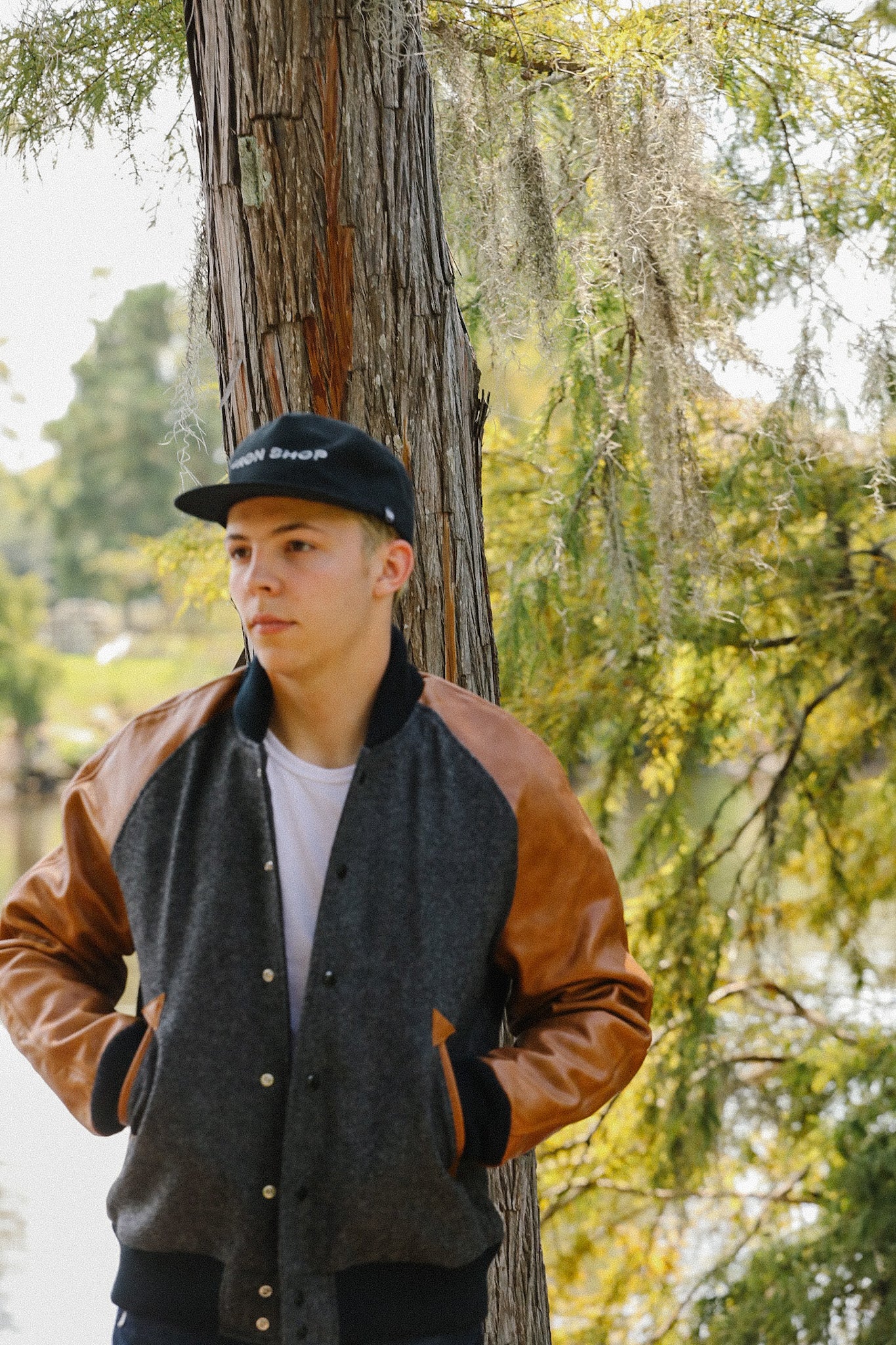 Young man wearing Iron Shop cap and leather-sleeve jacket standing outdoors by tree