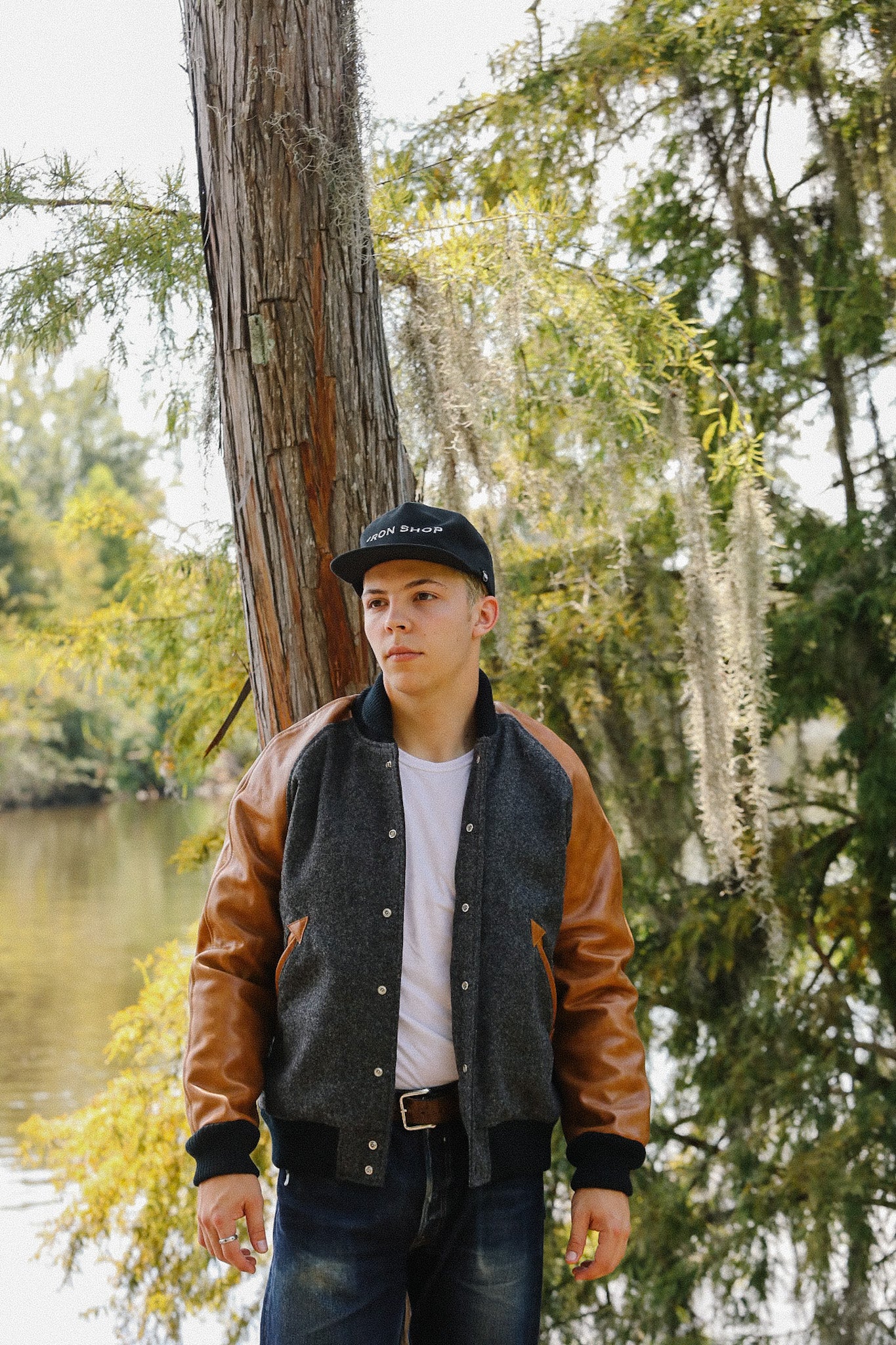 Young man wearing Iron Shop Provisions jacket and cap standing by a tree near a calm river