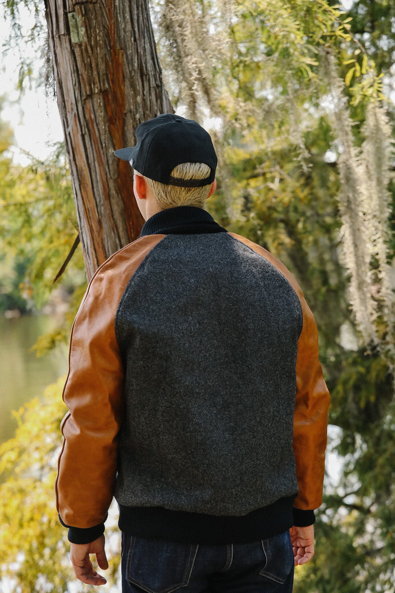 Man wearing black cap and brown leather sleeve wool jacket standing near tree in outdoor nature setting