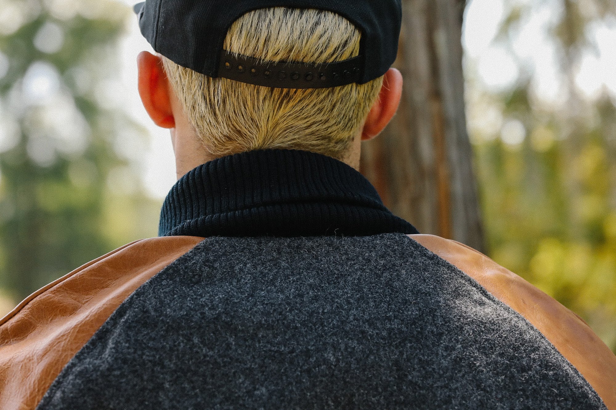 Back view of person with bleached blonde hair wearing black cap and gray jacket with brown leather sleeves outdoors