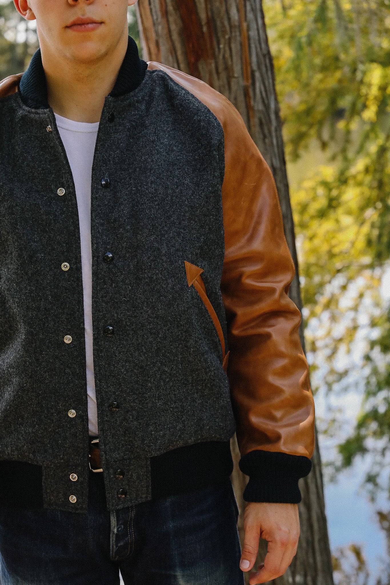 Man wearing grey wool varsity jacket with brown leather sleeves standing outdoors near tree