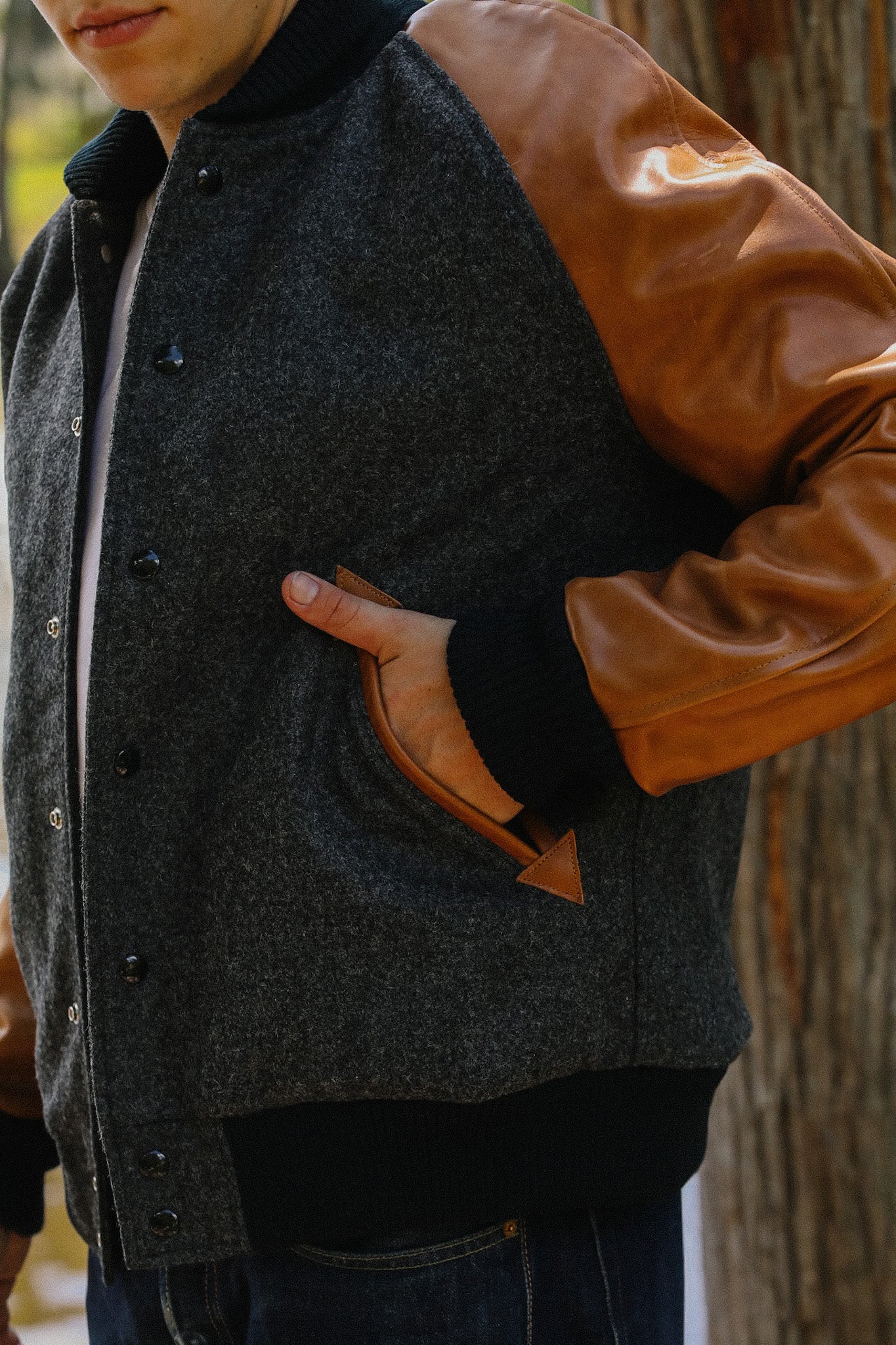 Close-up of a man wearing a dark grey wool varsity jacket with tan leather sleeves and black ribbed cuffs, outdoors