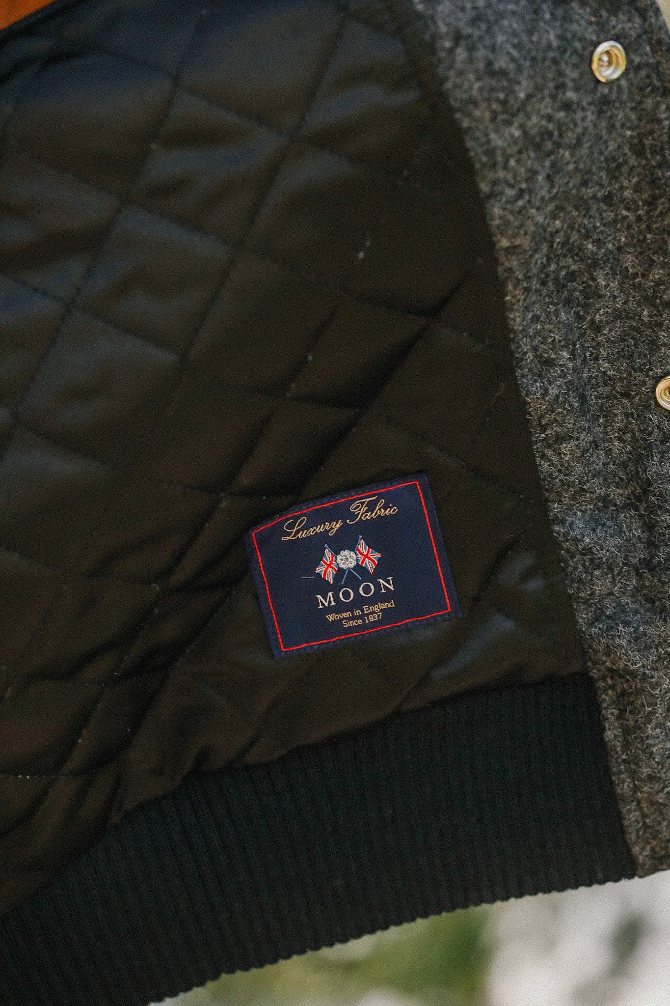 Close-up of luxury quilted fabric label reading MOON, woven in England since 1837, on black and grey textured jacket