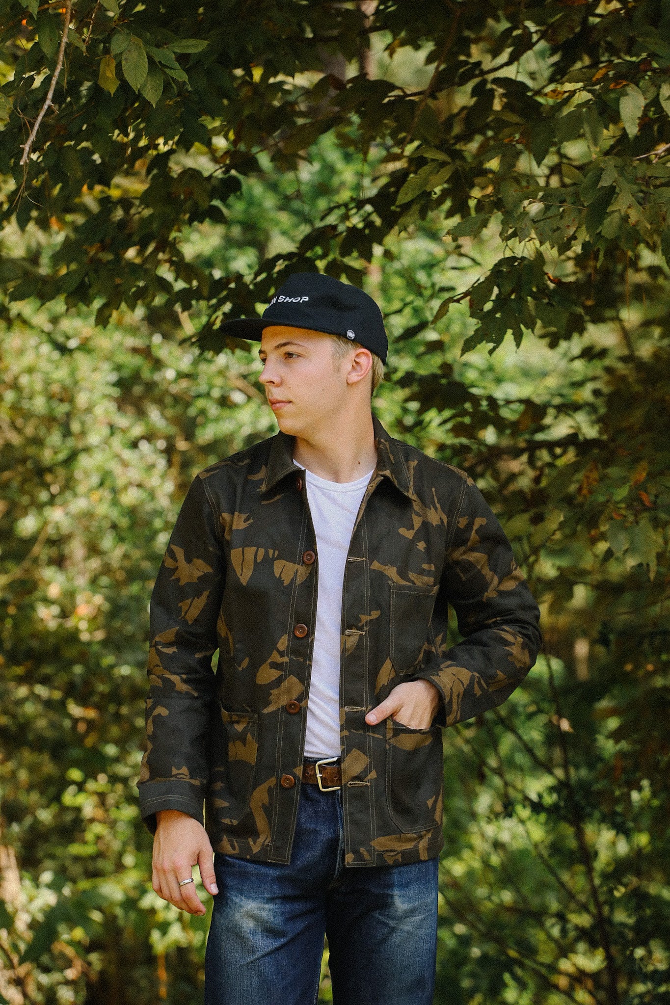 Young man in camo jacket and black Iron Shop cap standing outdoors with green tree background