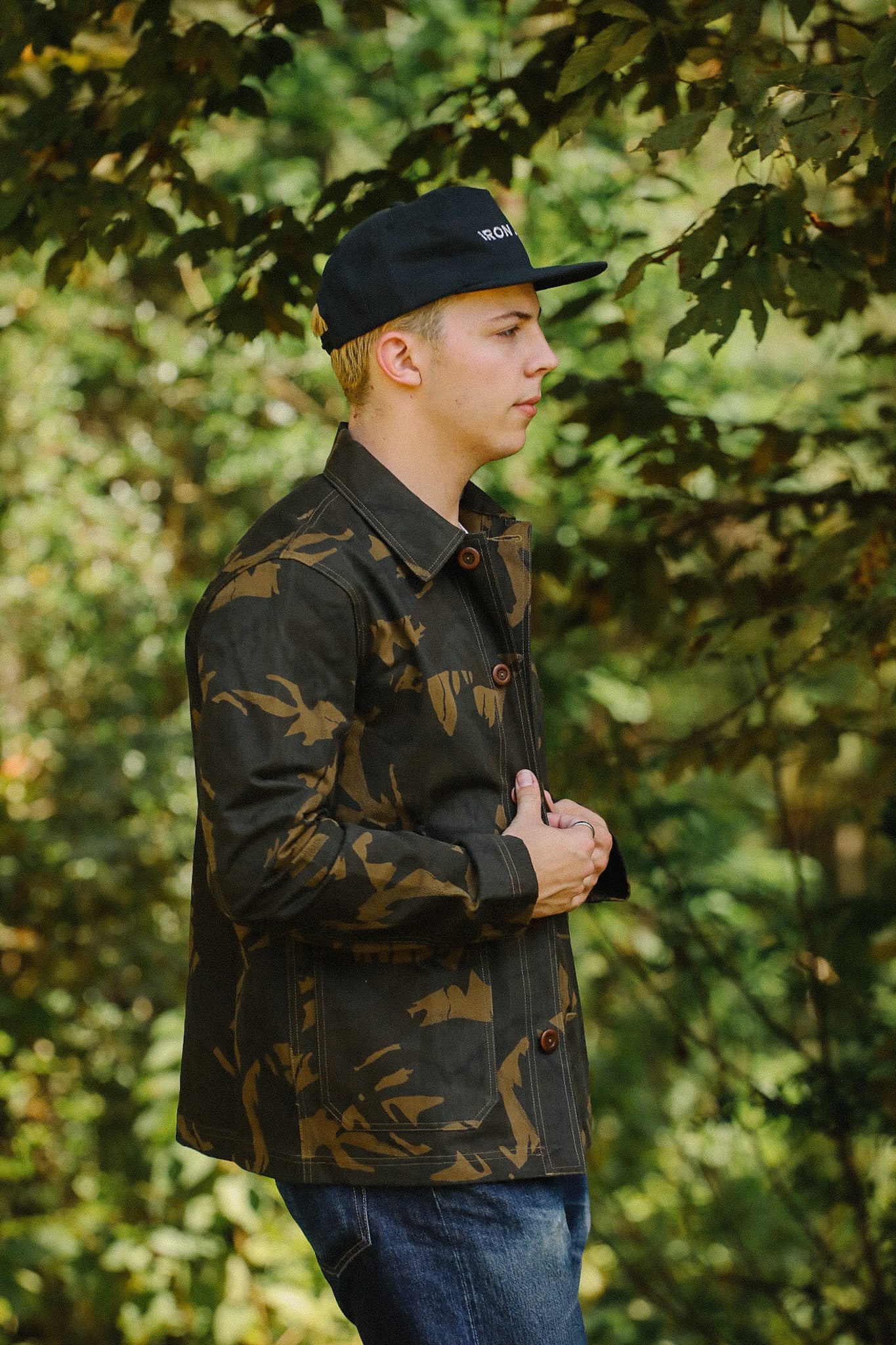 Side profile of a young man wearing a camo jacket and black Iron Shop Provisions cap in a leafy outdoor setting