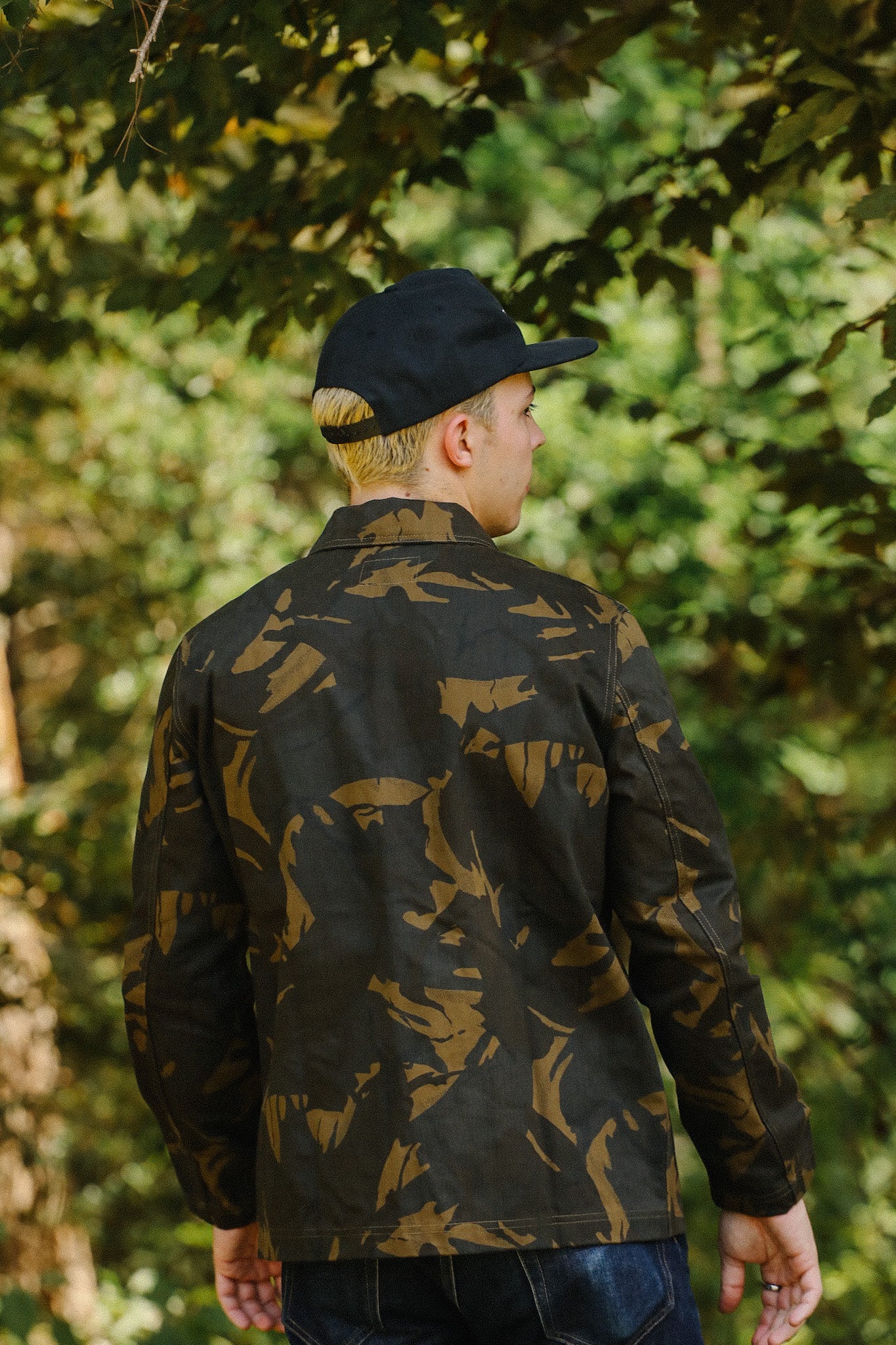 Young man wearing camo jacket and black cap standing outdoors with green foliage background