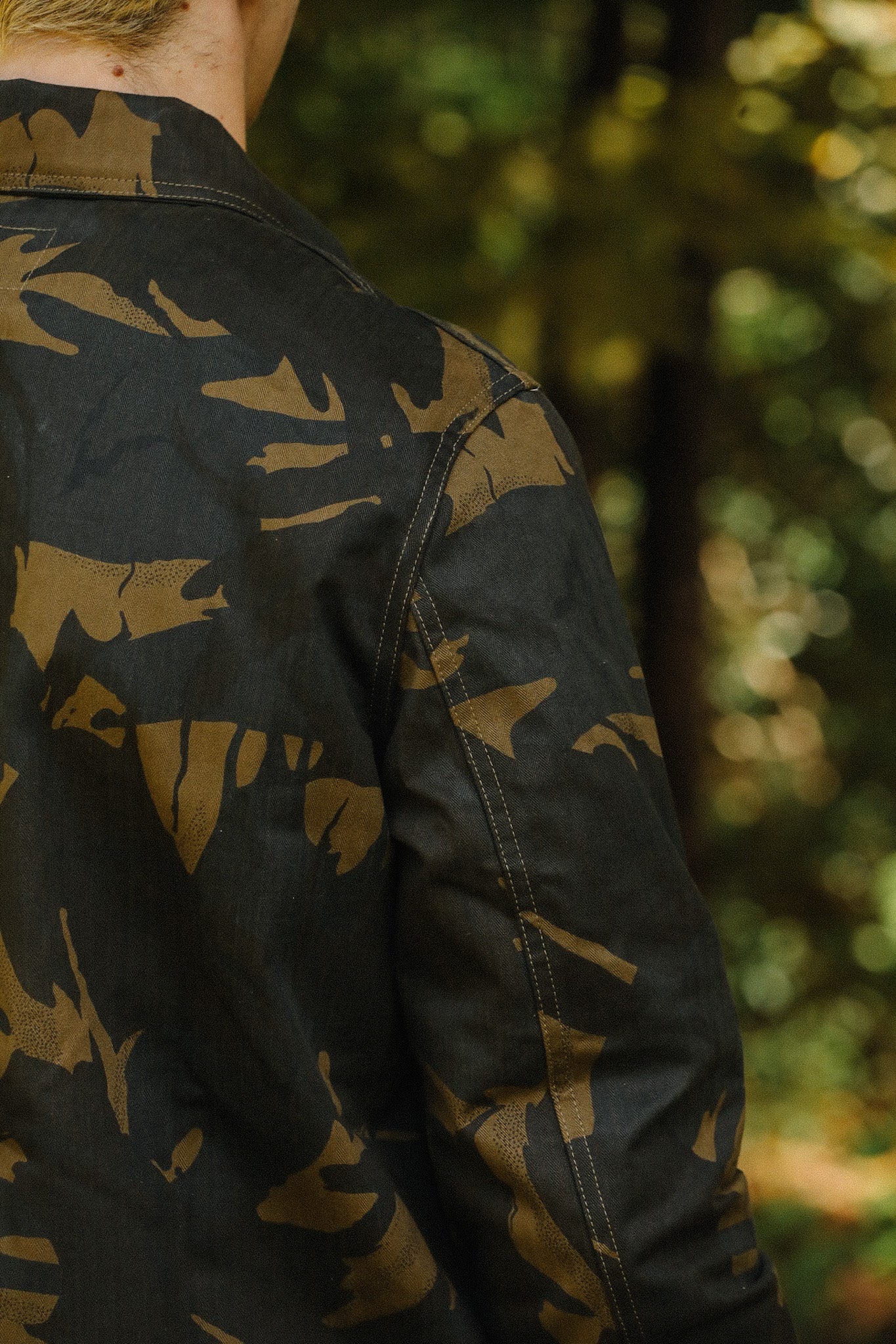 Close-up of a person wearing a black and brown camo jacket with visible stitching in an outdoor forest setting
