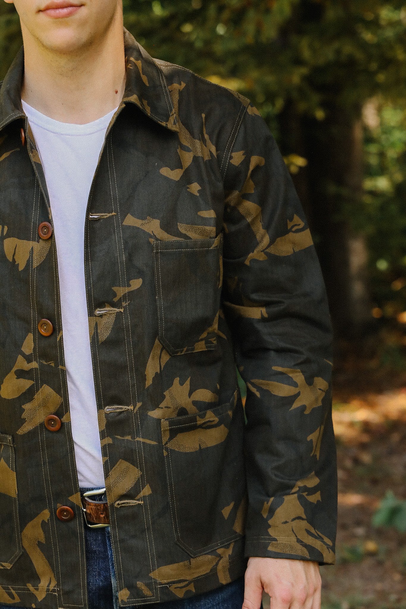 Man wearing a camouflage patterned jacket with brown buttons and pocket over a white t-shirt outdoors