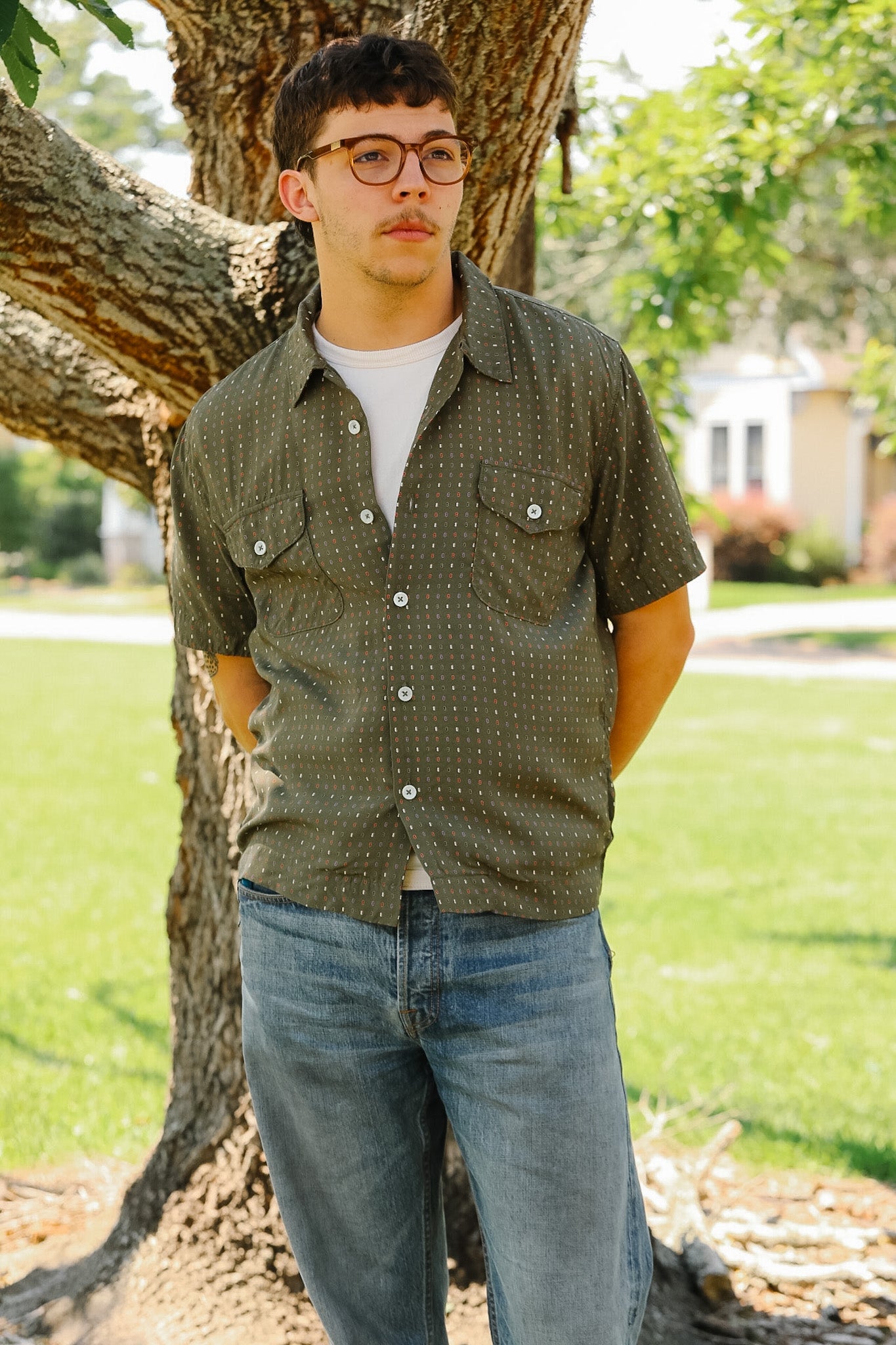 Man wearing a green patterned shirt and blue jeans standing next to a tree in a park.