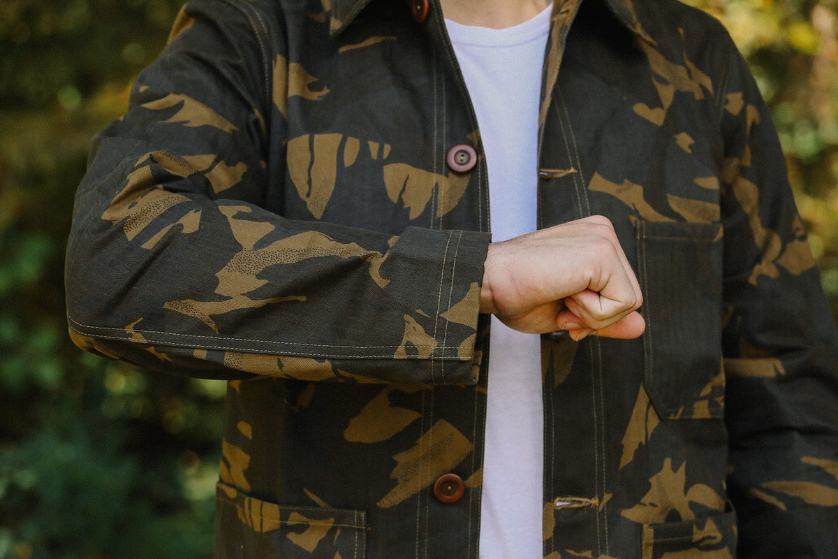 Person wearing camo jacket with wooden buttons and white shirt background outdoors