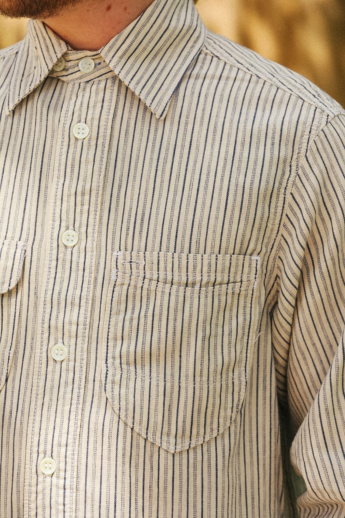Close-up of a man wearing a beige and navy vertically striped button-down shirt, showing the collar, front buttons, and chest pocket.