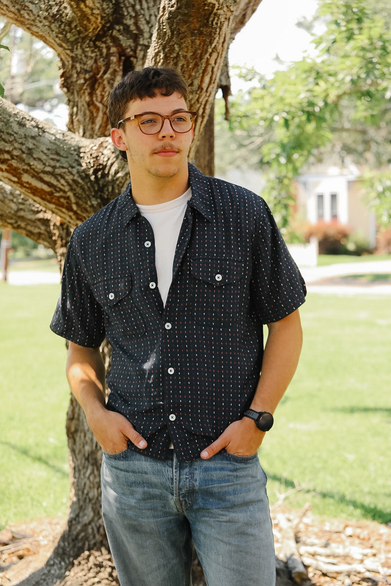 Man wearing a patterned shirt and jeans standing under a tree
