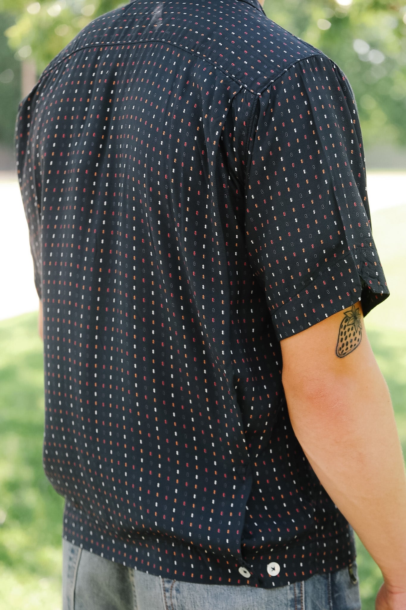Person wearing a black patterned shirt outdoors with greenery in the background