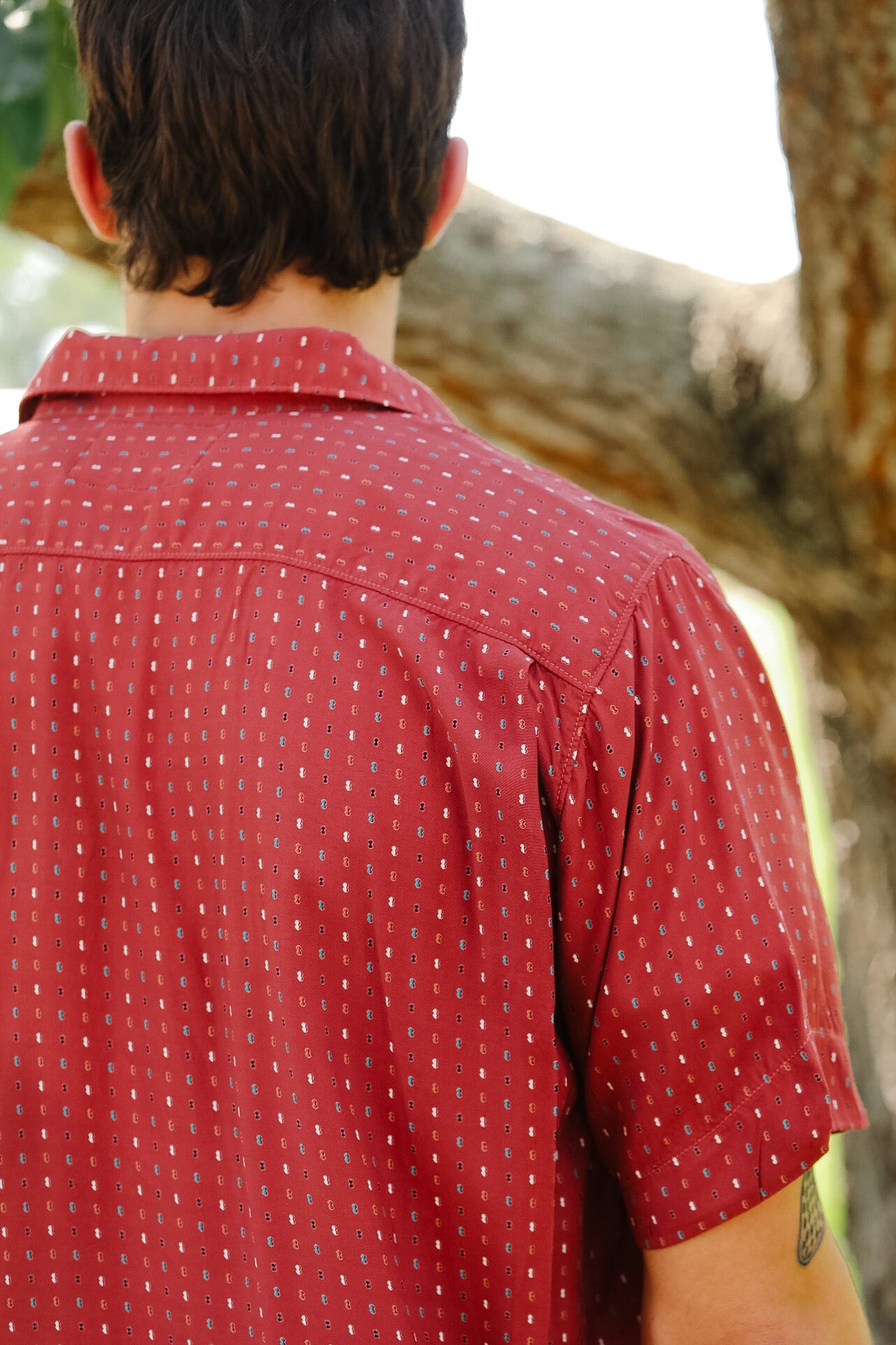 Person wearing a red patterned shirt standing outdoors with trees in the background