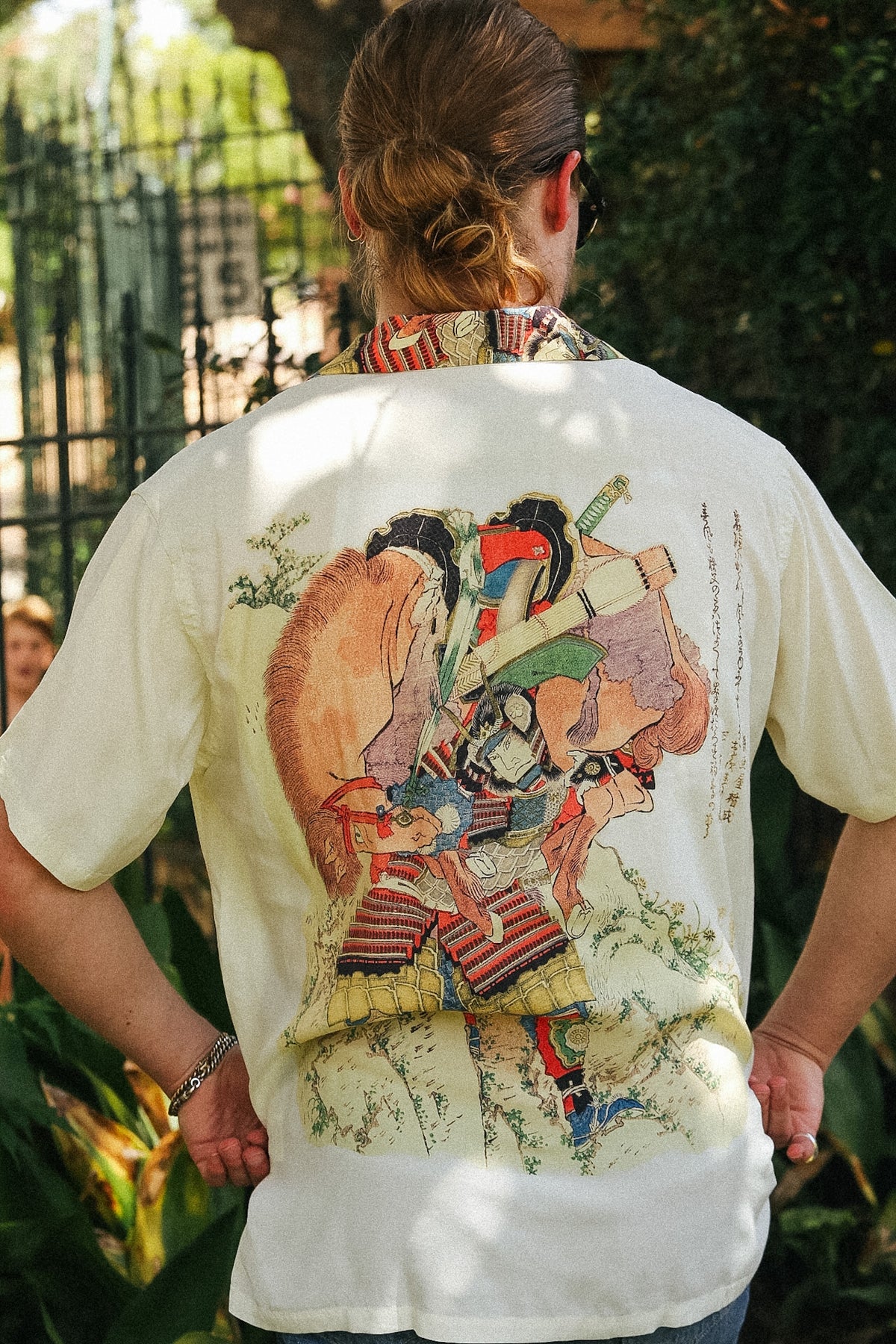 A person viewed from behind wearing a cream-colored short-sleeve t-shirt with a large, detailed Japanese ukiyo-e style graphic print on the back, set against an outdoor garden background.